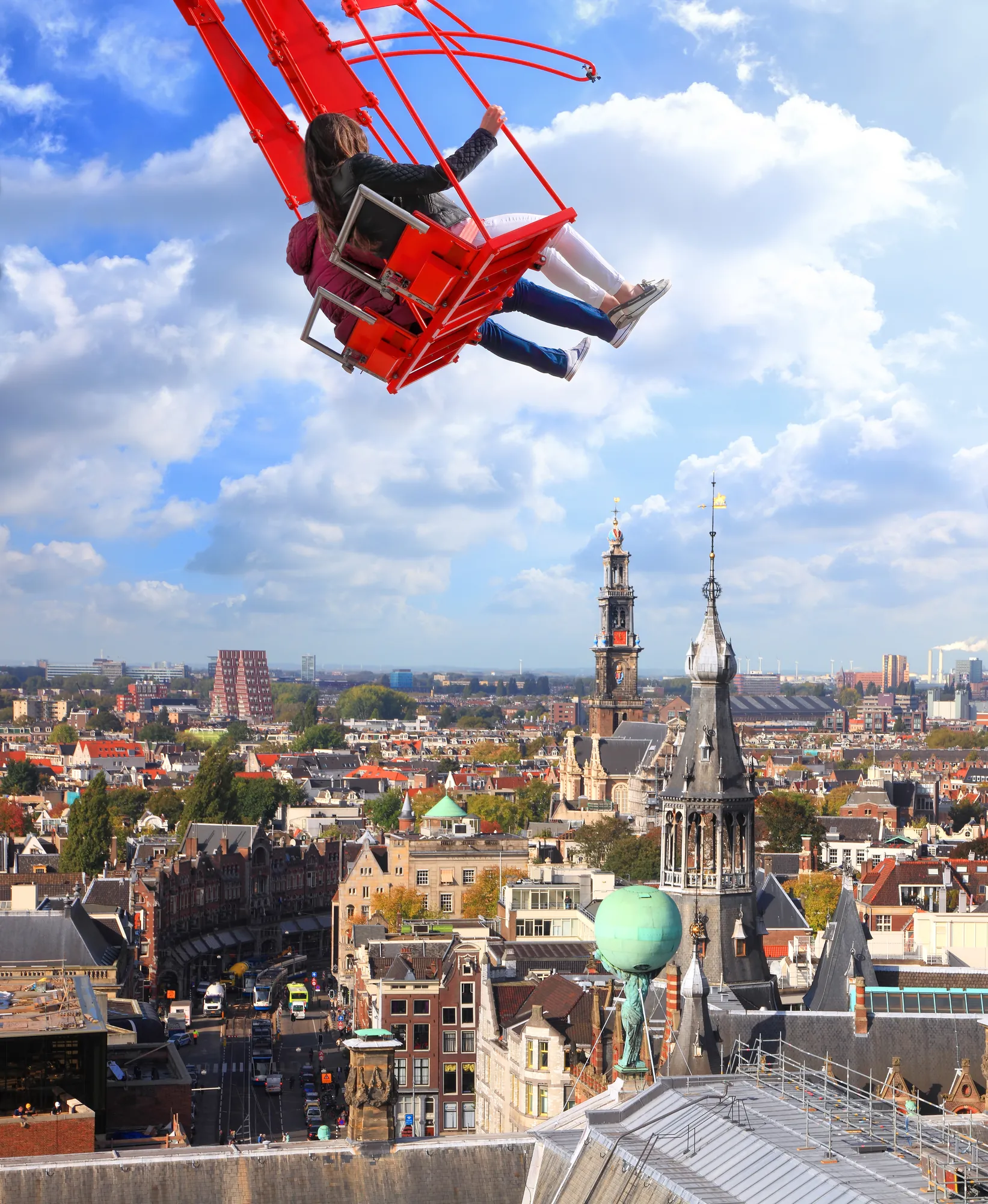 Pärchen in der A’DAM Lookout Schaukel, schwingend über den Dächern von Amsterdam bei Tageslicht, mit einem beeindruckenden Blick auf die Stadt und den klaren Himmel im Hintergrund.