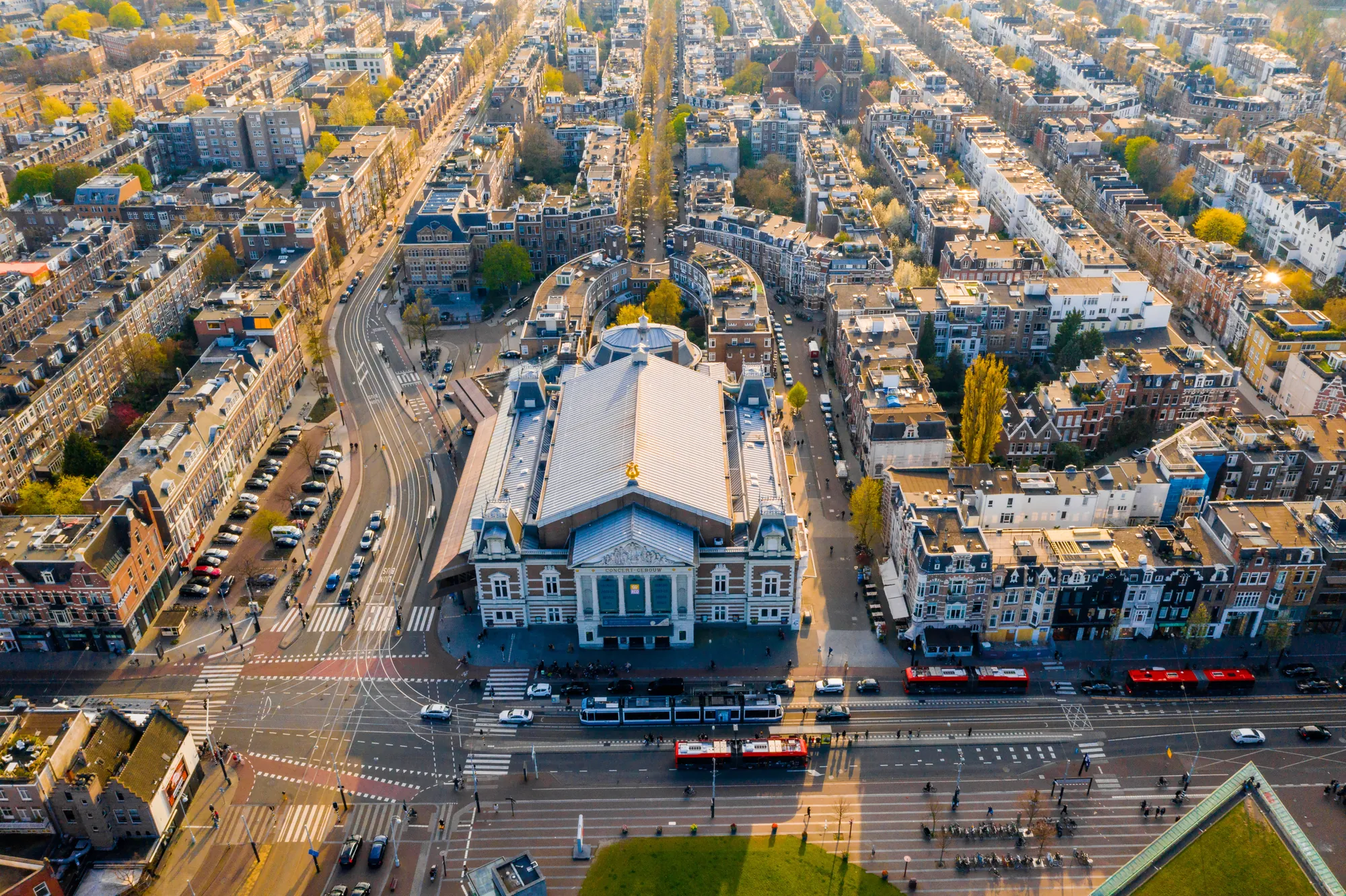 Panoramaansicht über das Concertgebouw in Amsterdam, umgeben von gepflegten Grünflächen und eleganten Stadtgebäuden, bei strahlendem Sonnenlicht, das die architektonischen Details hervorhebt.