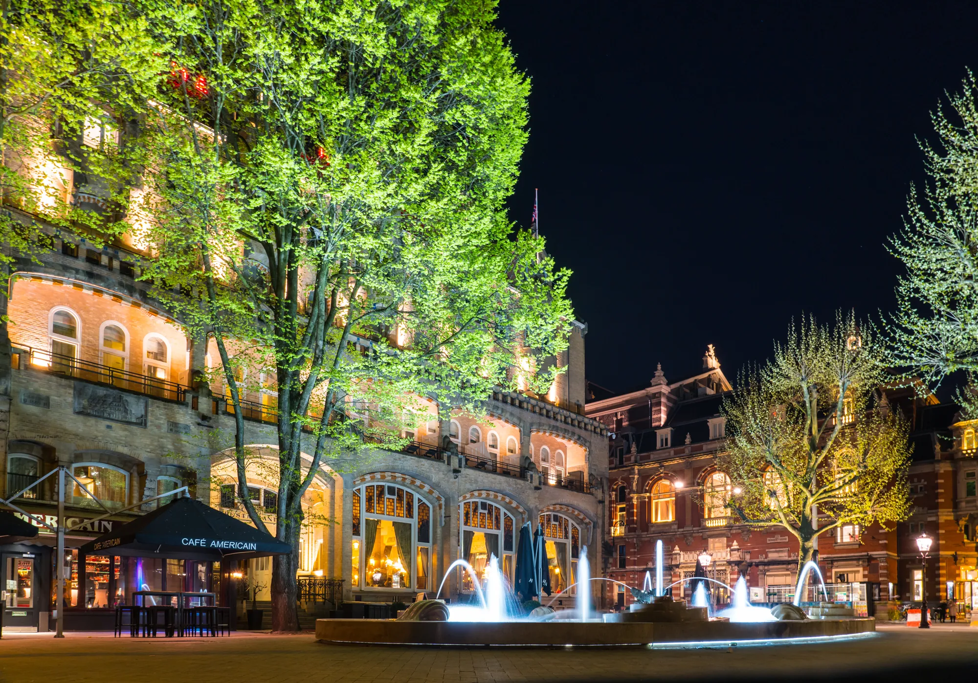 Das Café Americain am Leidseplein in der Nacht, elegant beleuchtet, mit einem funkelnden Brunnen im Vordergrund. Das Licht des Cafés und des Brunnens spiegelt sich auf dem Platz und erzeugt eine ruhige, aber eindrucksvolle Szenerie.
