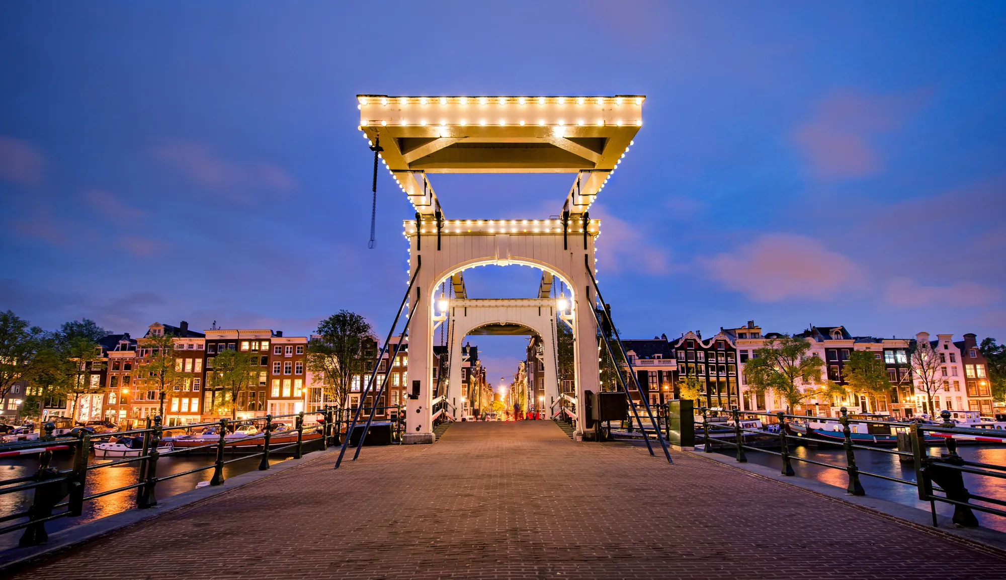 Das Bild zeigt die Magere Brug in Amsterdam während der Abenddämmerung. Die Brücke ist festlich beleuchtet, und die Lichter spiegeln sich im ruhigen Wasser des Kanals. Im Hintergrund sind typische Amsterdamer Giebelhäuser zu sehen, deren Fenster warm erleuchtet sind, was eine gemütliche Atmosphäre schafft.