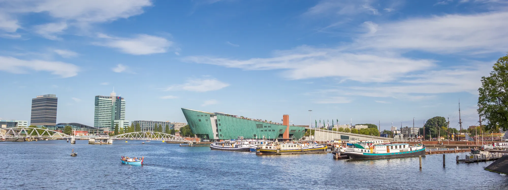 Das Bild zeigt eine Panoramaansicht des NEMO Science Museums in Amsterdam bei klarem Wetter. Im Vordergrund befindet sich das Wasser mit verschiedenen Booten und das markante kupfergrüne Gebäude des Museums erhebt sich imposant vor einem strahlend blauen Himmel. Im Hintergrund sind moderne Gebäude und eine Brücke zu sehen, die die urbane Skyline der Stadt prägen.