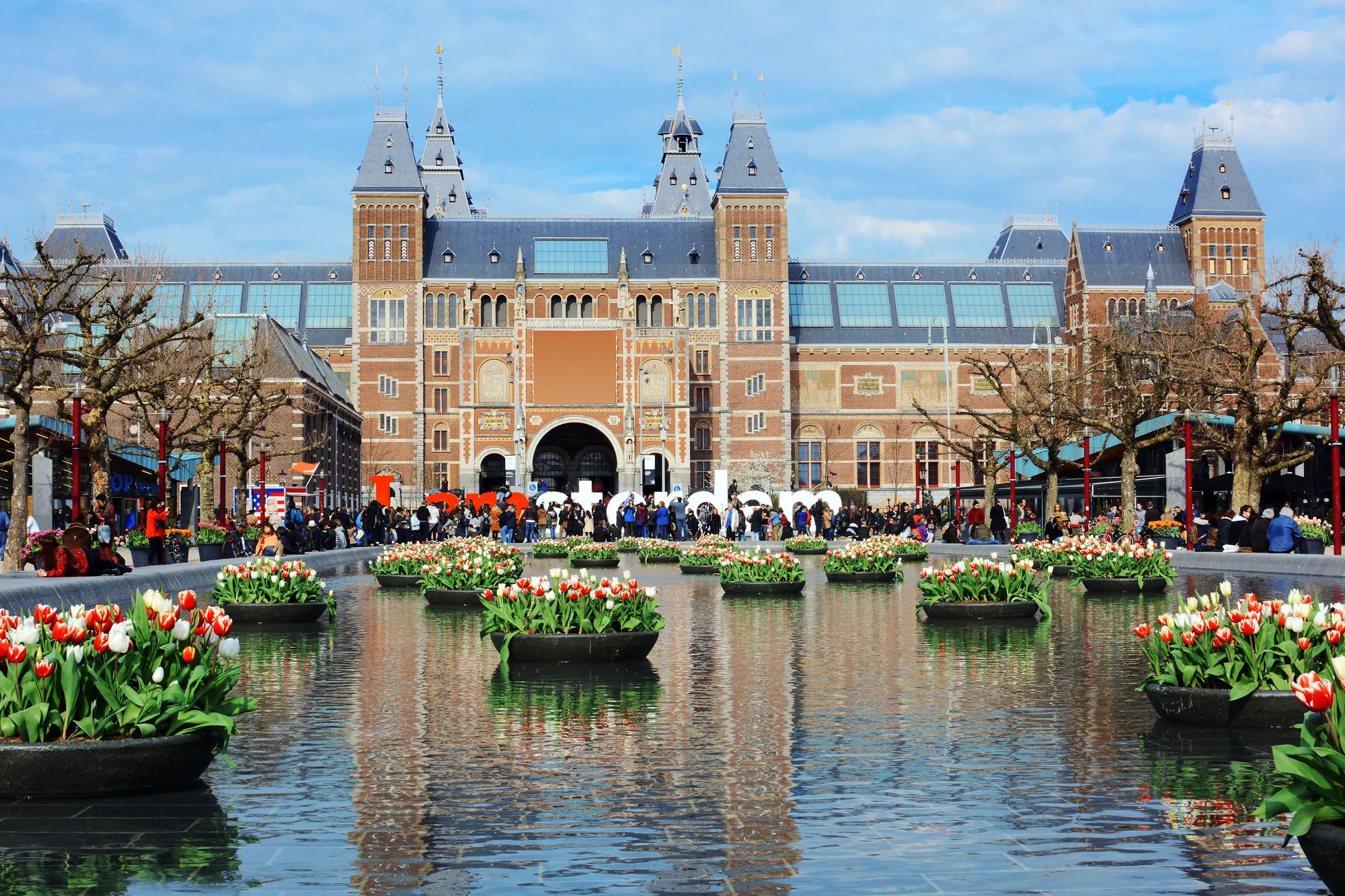 Farbenfrohe Tulpenschalen schwimmen auf dem Wasser vor dem Rijksmuseum in Amsterdam, umgeben von zahlreichen Besuchern, die das fröhliche Treiben genießen. Die beeindruckende Architektur des Museums bildet einen schönen Kontrast zu den lebhaften Blumen und dem lebhaften Ambiente der Umgebung.