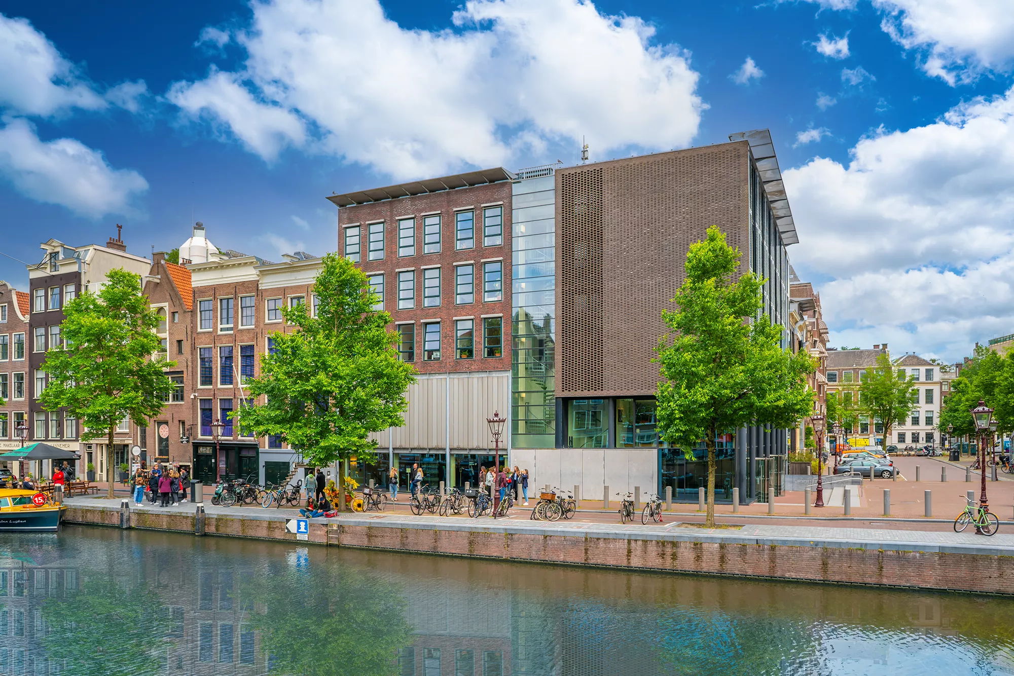 Sommerlicher Blick auf das Anne Frank Haus vom Kanal in Amsterdam, umgeben von üppigem Grün und blühenden Blumen. Die lebhafte Atmosphäre wird durch die vorbeifahrenden Boote und die farbenfrohen Fassaden der umliegenden Gebäude ergänzt.