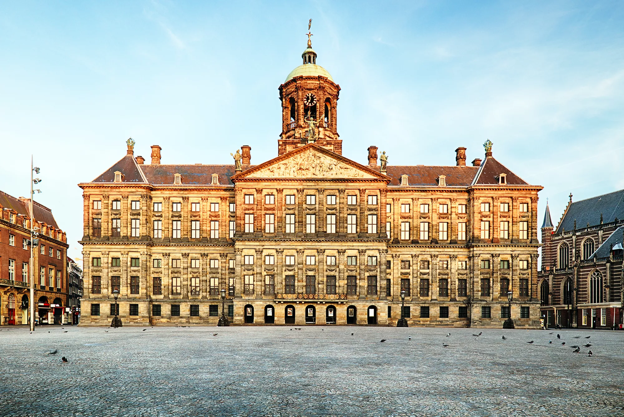 Königspalast in Amsterdam mit prunkvoller Fassade im Herzen der Stadt, umgeben von Tauben, die vor dem Gebäude verweilen. Der klare Himmel hebt die architektonische Schönheit hervor und verleiht der Szene eine lebendige Atmosphäre.