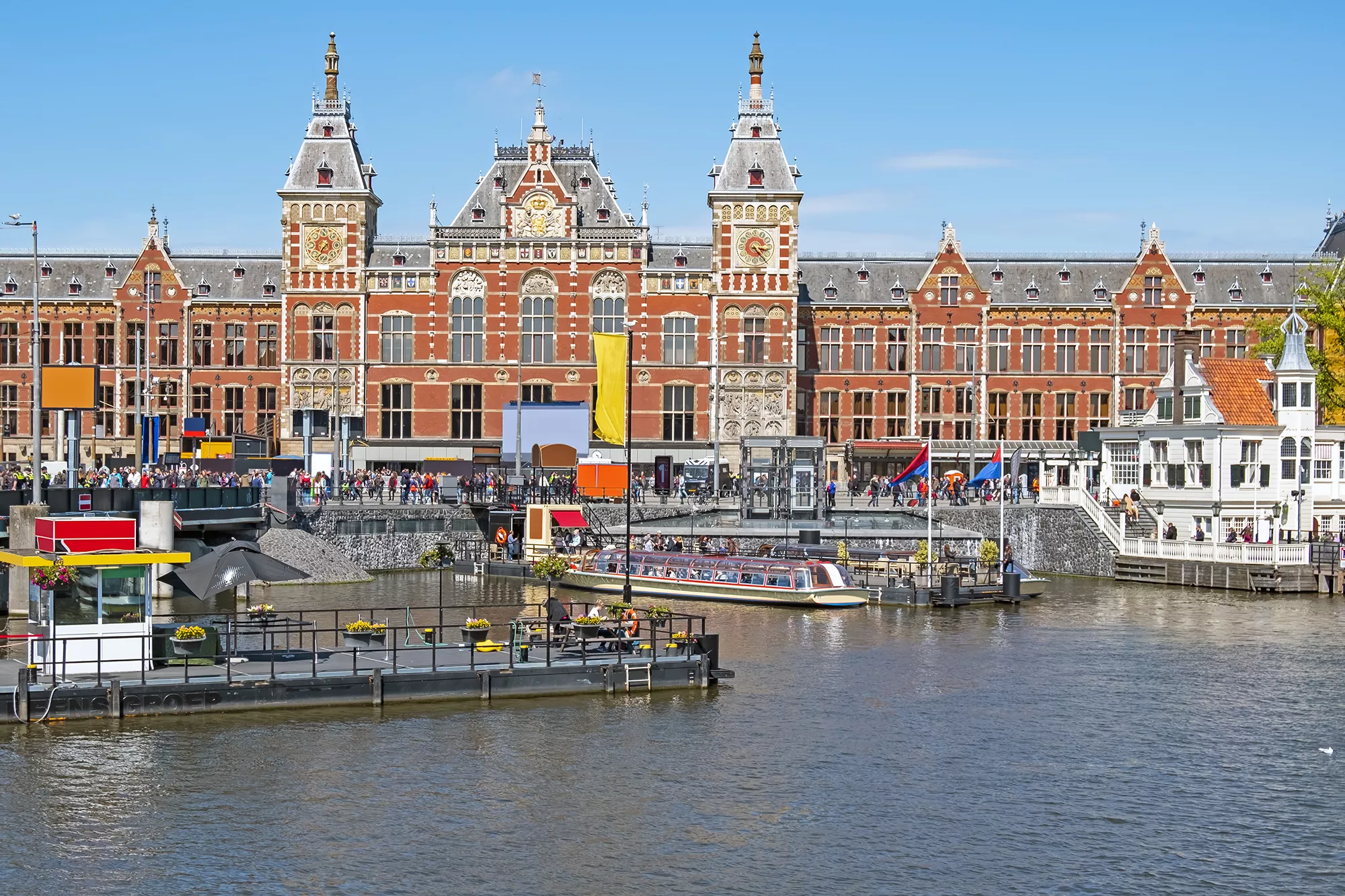 Bahnhof in Amsterdam mit Anlegestelle im Vordergrund, bei sonnigem Wetter. Die Architektur des Gebäudes ist gut sichtbar, während Boote sanft im Wasser schaukeln und die Umgebung lebendig wirkt.