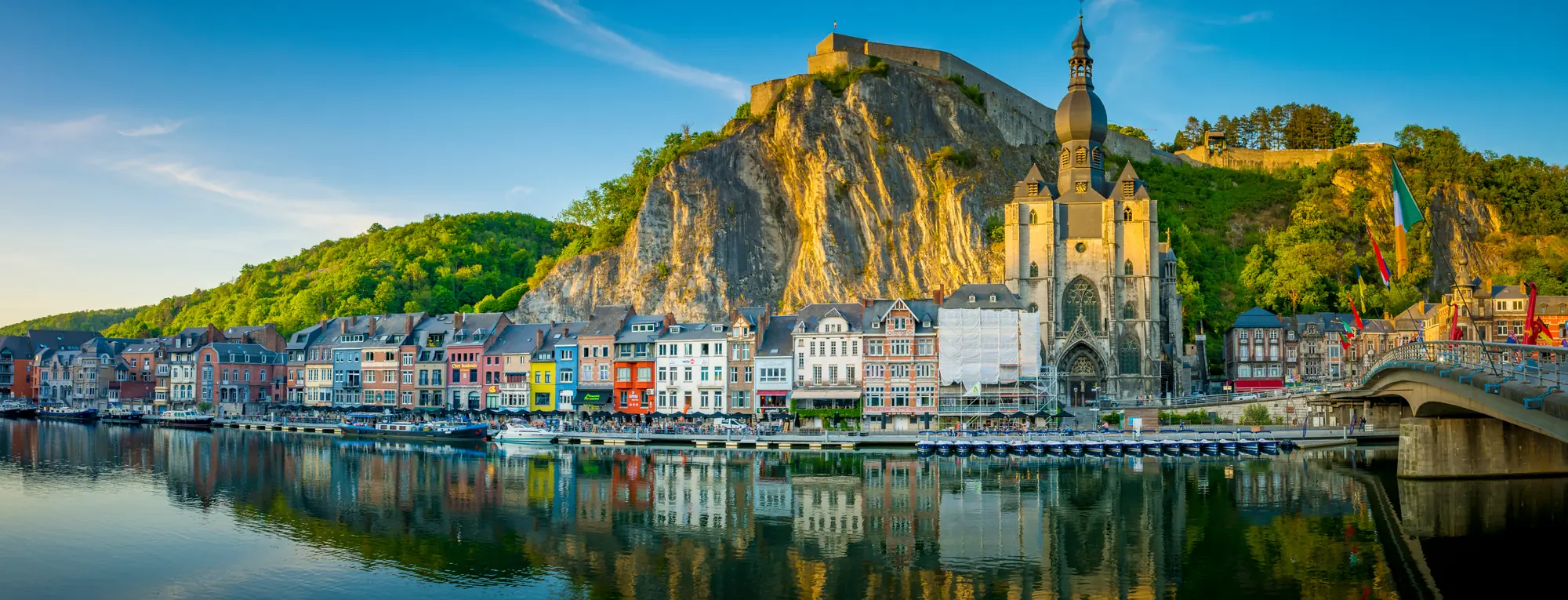 Blick vom Flussufer der Maas auf Dinant in den Ardennen bei sonnigem, klarem Himmel. Die Stadt, umrahmt von steilen Klippen, zeigt die jahrhundertealte Zitadelle, die majestätisch auf einem Felsen oberhalb thront.