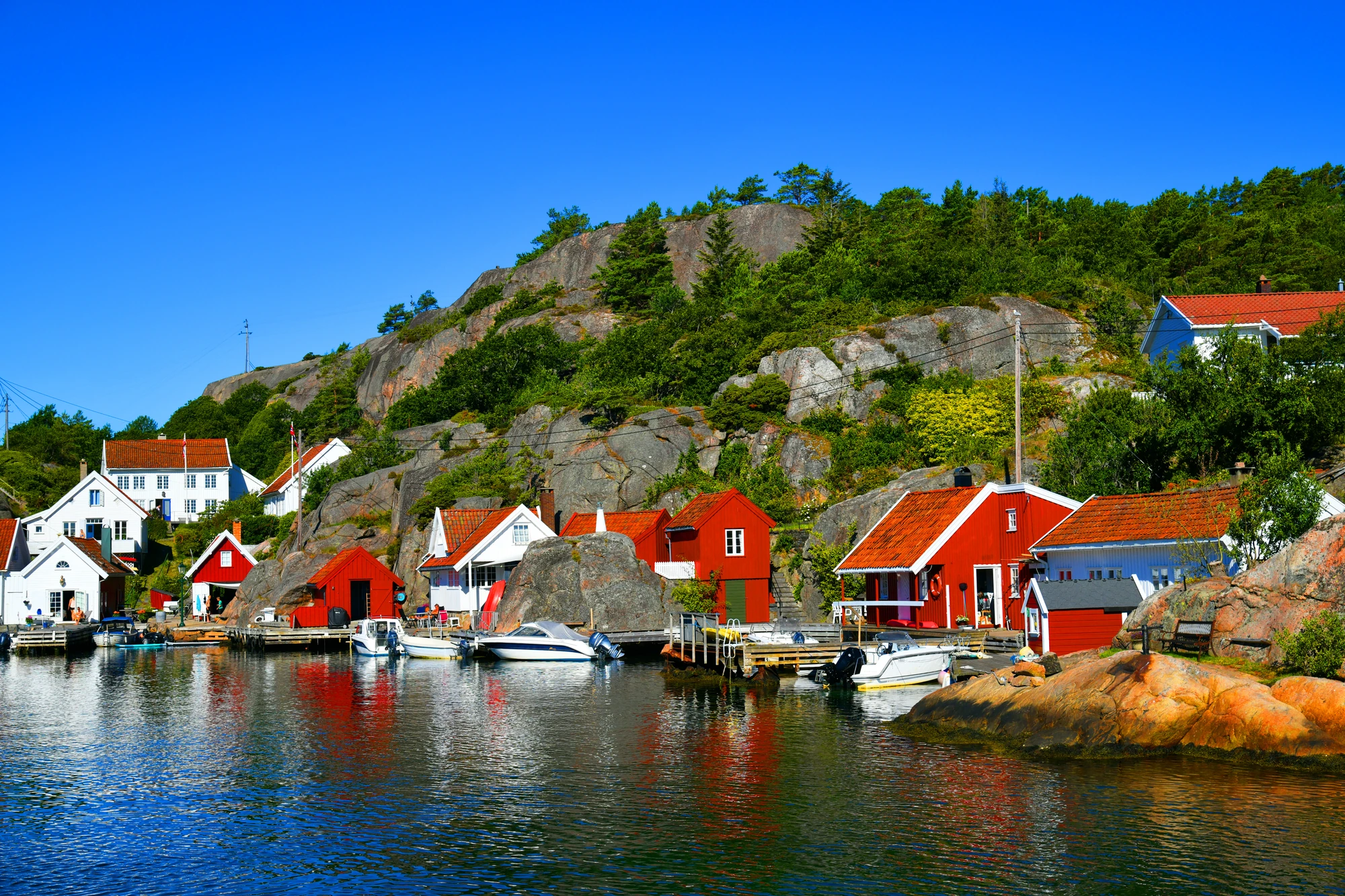 Kleine rote und weiße Häuser am Wasser mit Booten, dahinter bewaldeter Felsen unter blauem Himmel in Kristiansand in Norwegen