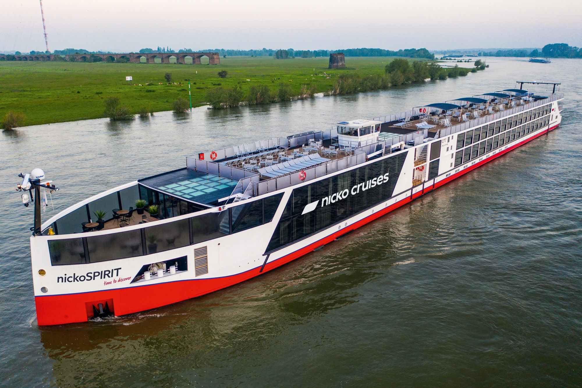 Blick am Abend auf die Außenansicht der nickoSPIRIT bei Fahrt auf einem Fluss vorbei an Feldern mit einer Brücke im Hintergrund