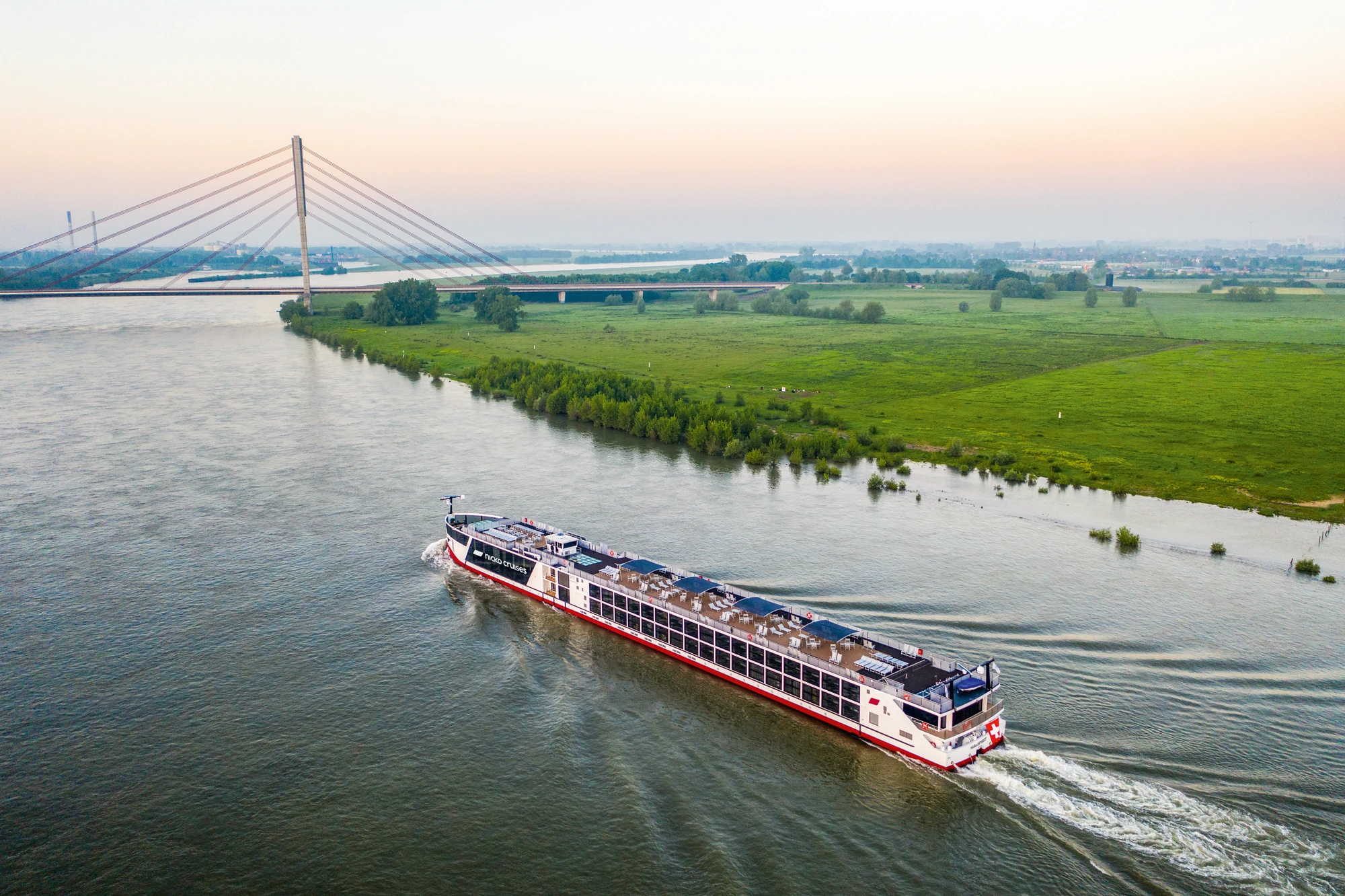 Blick am Abend auf die Außenansicht der nickoSPIRIT bei Fahrt auf einem Fluss vorbei an Feldern mit einer Stadt und einer Brücke im Hintergrund