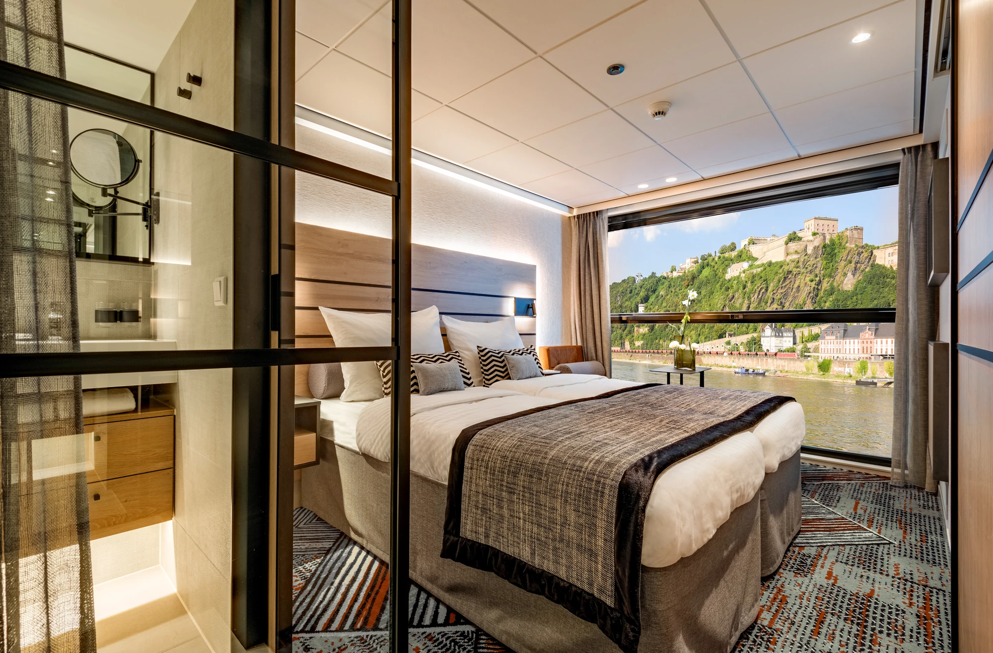 Kabine auf dem Mittel- oder Oberdeck mit einem Doppelbett und Panoramafenstern im Hintergrund und einem Blick in das Badezimmer