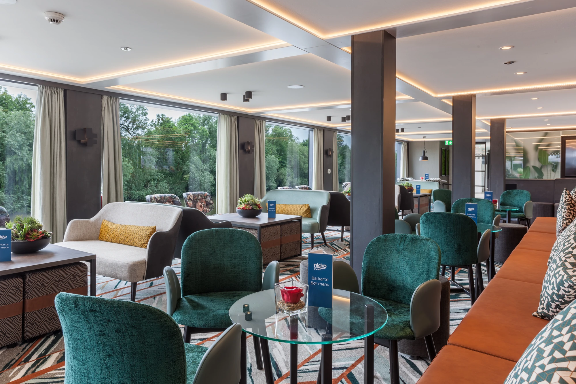 Bunte Stühle und Sitzbänke vor gläsernen Rundtischen mit Pflanzen und Kerzen im Salon an Bord der nickoSPIRIT