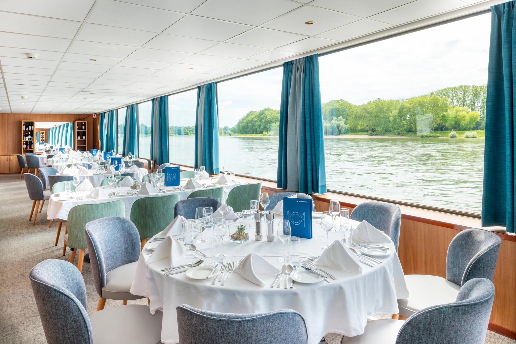 Gedeckte Tische und Stühle im Restaurant der RHEIN MELODIE mit großen Panoramafenstern bei Fahrt auf einem Fluss