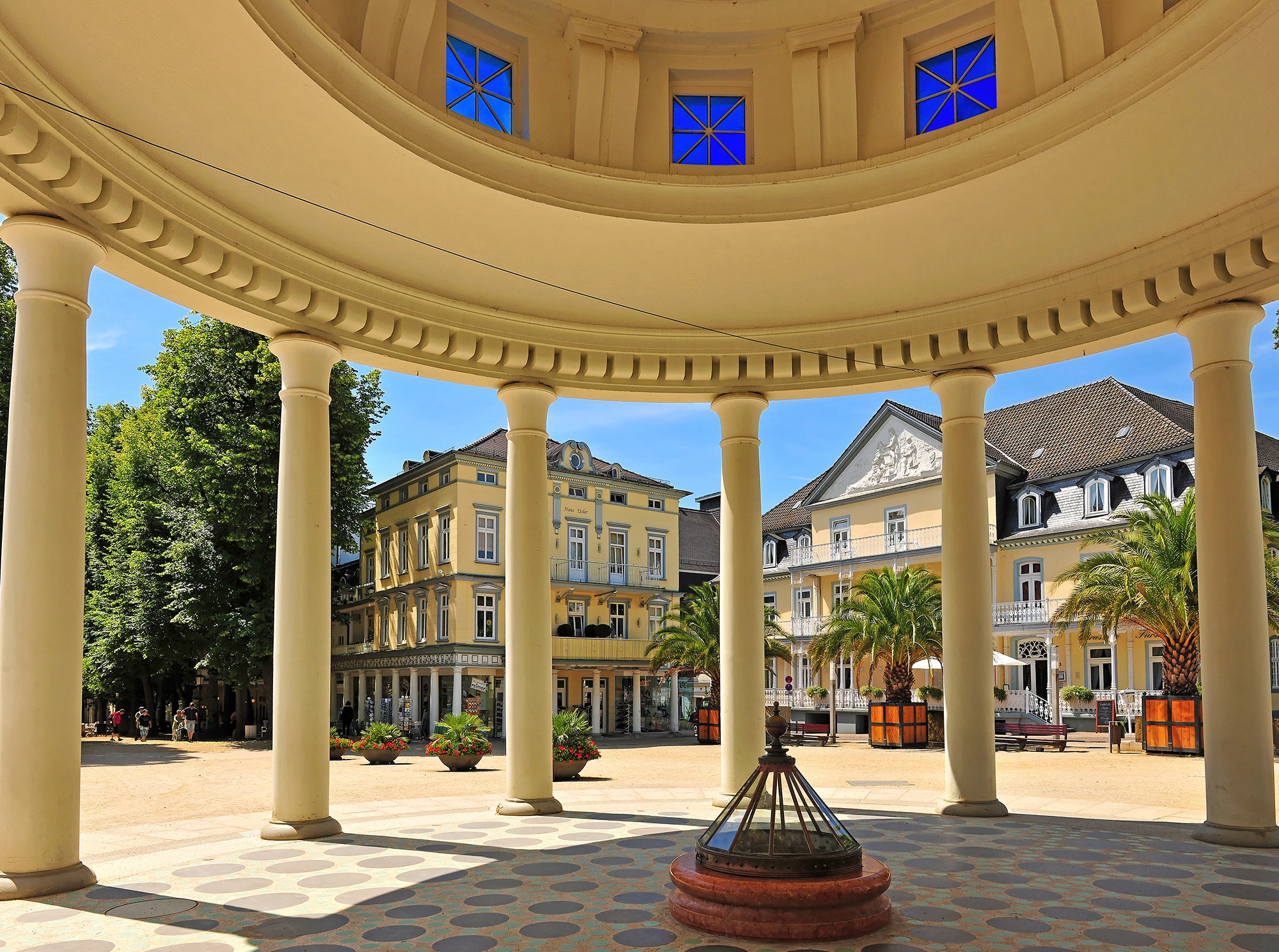 Das Bild zeigt einen charmanten Blick auf den historischen Kurpark von Bad Pyrmont, eingefangen durch die elegante Säulenstruktur eines Pavillons. Im Hintergrund erheben sich prächtige, klassizistische Gebäude, die durch ihre detailreichen Fassaden und großen Fenster bestechen. Der gepflegte Platz, umgeben von Palmen und grünen Bäumen, vermittelt ein Gefühl von Ruhe und Erholung, typisch für die Atmosphäre eines Kurortes. Die harmonische Architektur und das sonnige Wetter verleihen dem Bild eine einladende und malerische Stimmung.