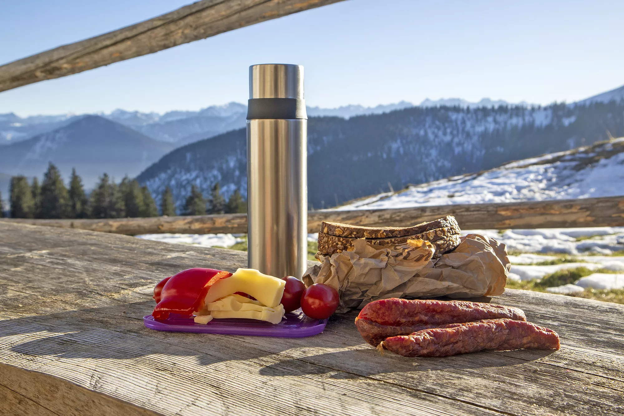 Bayerische Brotzeit mit Brot, Käse, Tomaten und Mettwürsten vor verschneiter Bergkulisse