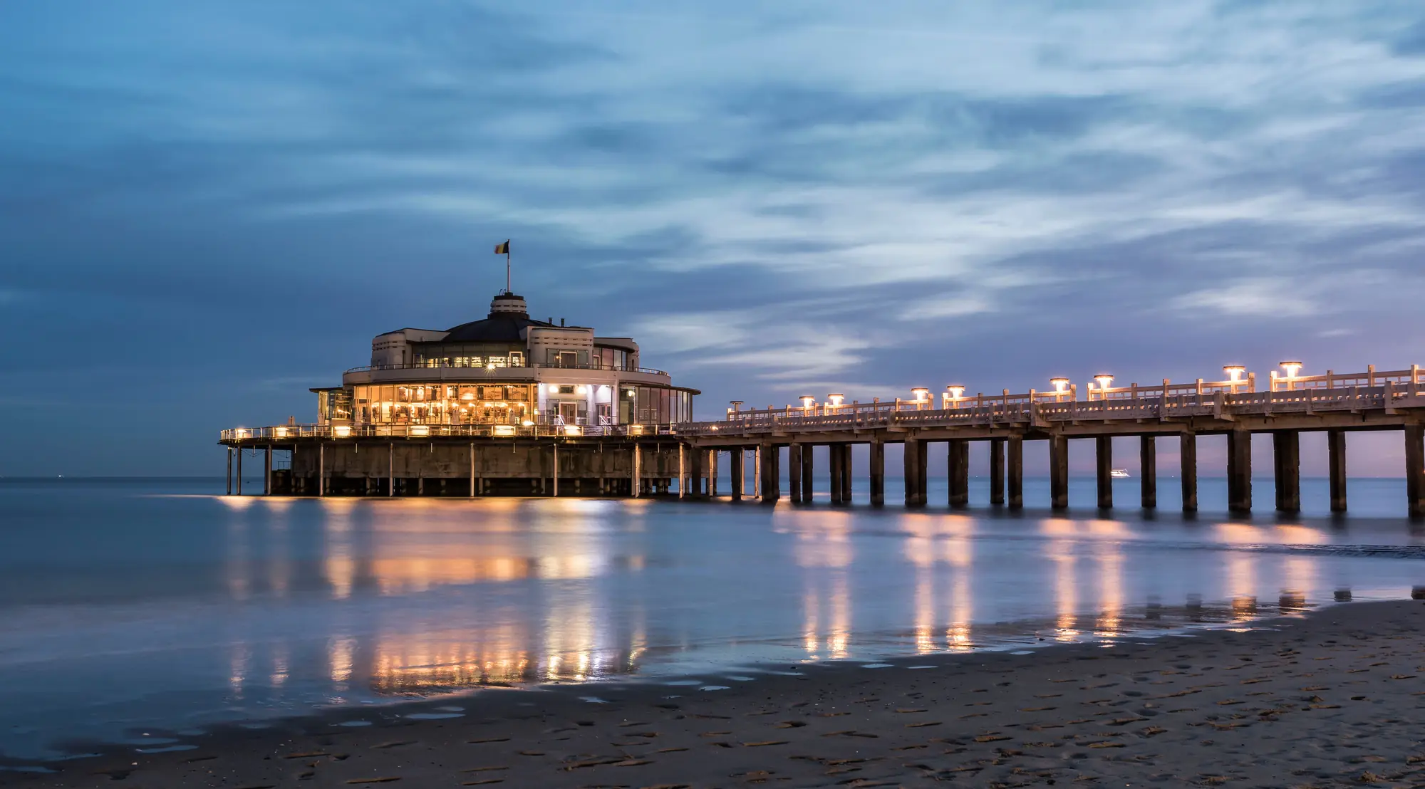 Beleuchteter belgischer Pier in der Abenddämmerung, der warmes Licht ausstrahlt und eine einladende Atmosphäre am Wasser schafft, umgeben von sanften Wellen und dem schwindenden Tageslicht.