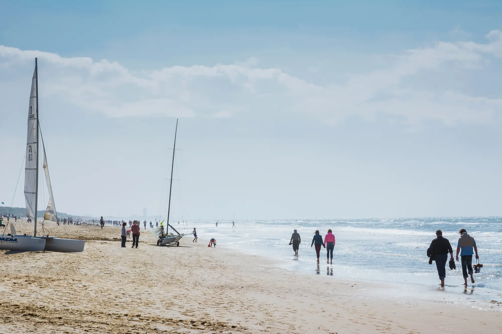 De Haan am Strand, wo Menschen am Ufer barfuß und im Sand spazieren. Der Himmel ist leicht bewölkt, während das Meer in der sanften Brise glitzert.