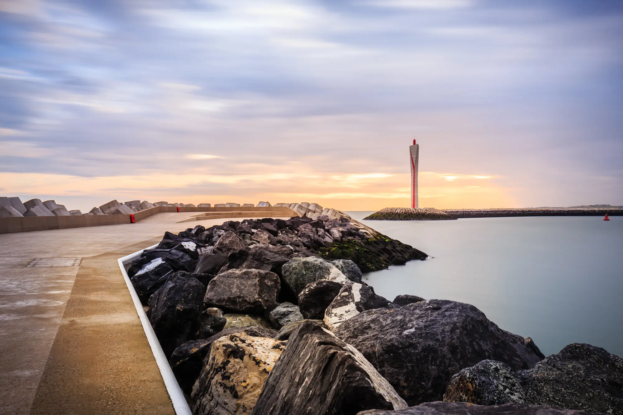 Oostende am Nieuwpoort Lighthouse bei Abenddämmerung, mit sanften Lichtreflexen auf dem Wasser und einer ruhigen Atmosphäre, die den historischen Leuchtturm und die umgebenden Steinfelsen hervorhebt.
