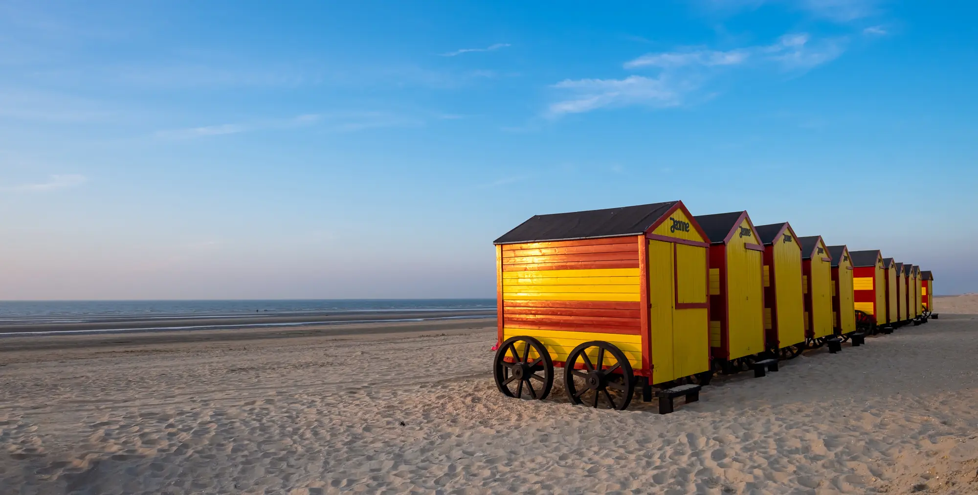 Gelb-rote Strandhütten auf Holzrädern an der belgischen Küste, die in der Sonne leuchten und eine lebendige Strandatmosphäre schaffen.