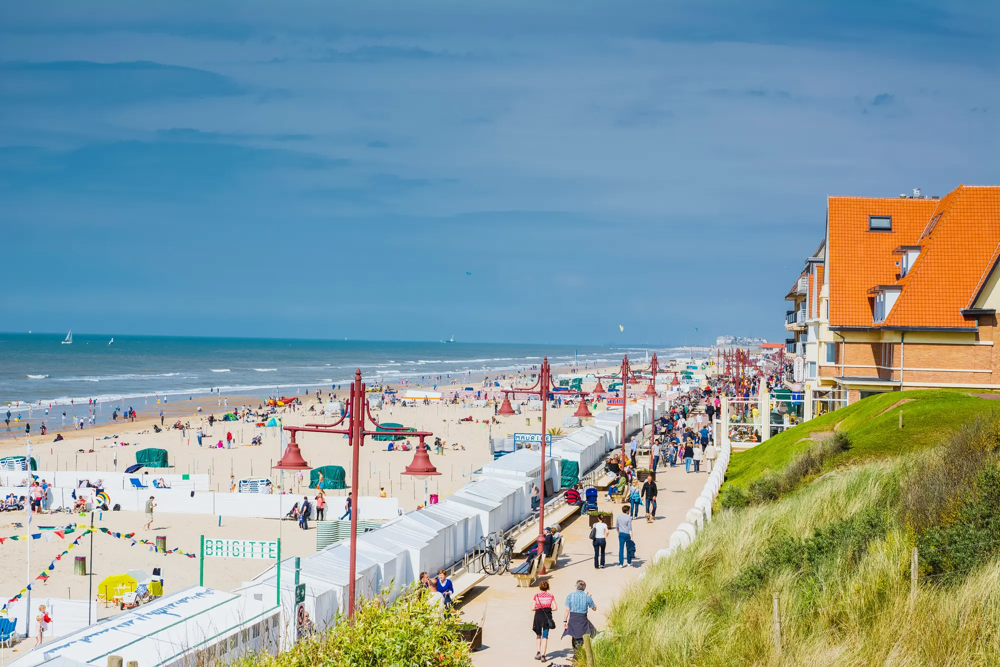 Gut besuchte Strandpromenade von De Haan in Belgien, mit Blick auf den Strand und das Meer bei milden Temperaturen, umgeben von fröhlichen Spaziergängern und bunten Strandkörben.
