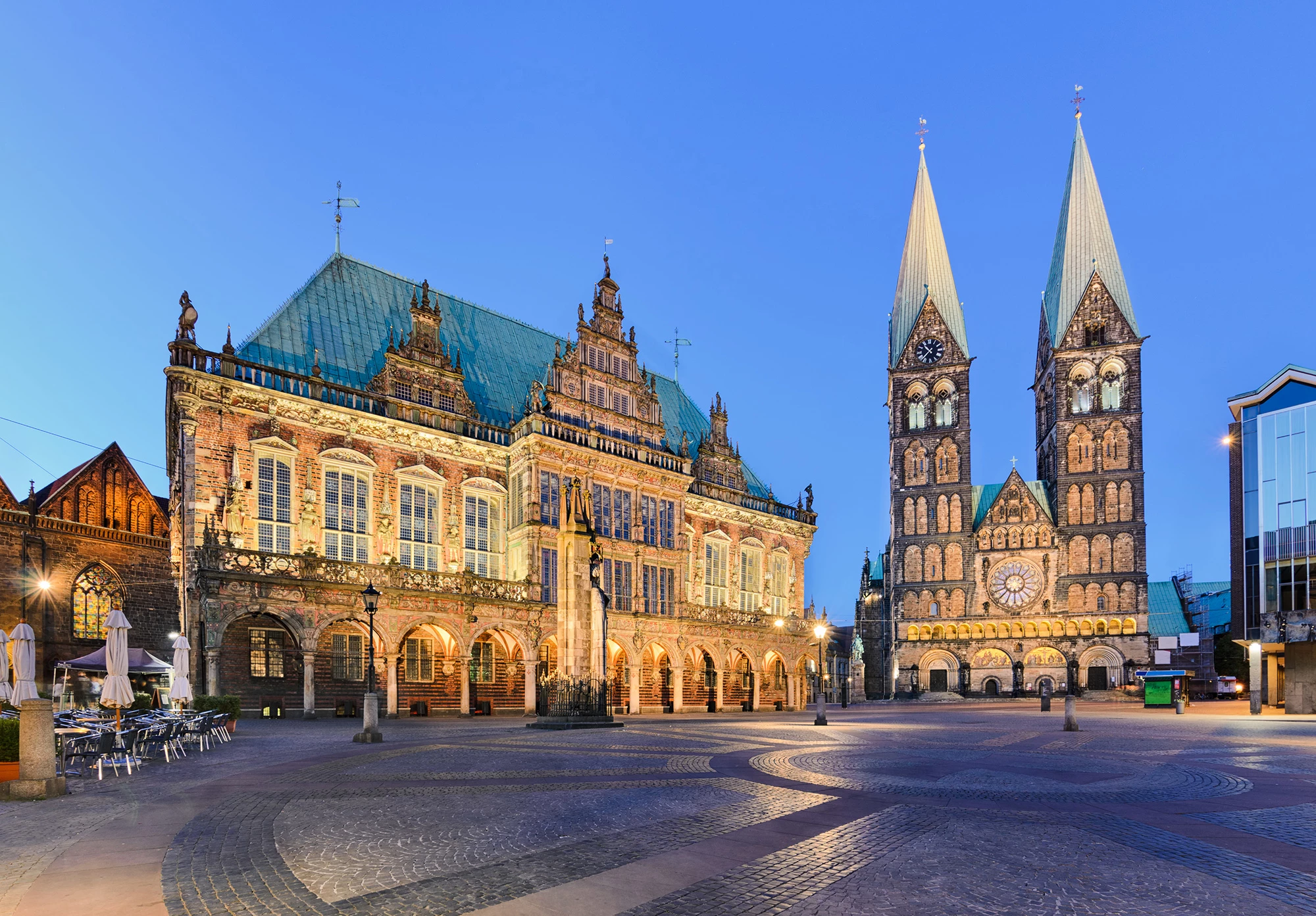 Der Bremer Marktplatz bei Abenddämmerung zeigt das prachtvolle, beleuchtete Bremer Rathaus mit seiner reich verzierten Fassade und den imposanten Dom St. Petri im Hintergrund. Der historische Platz wirkt ruhig, mit Kopfsteinpflaster und wenigen Menschen, während die Architektur im warmen Licht erstrahlt und die mittelalterliche Atmosphäre betont. Ein beeindruckendes Zusammenspiel von Kultur und Geschichte im Herzen Bremens.