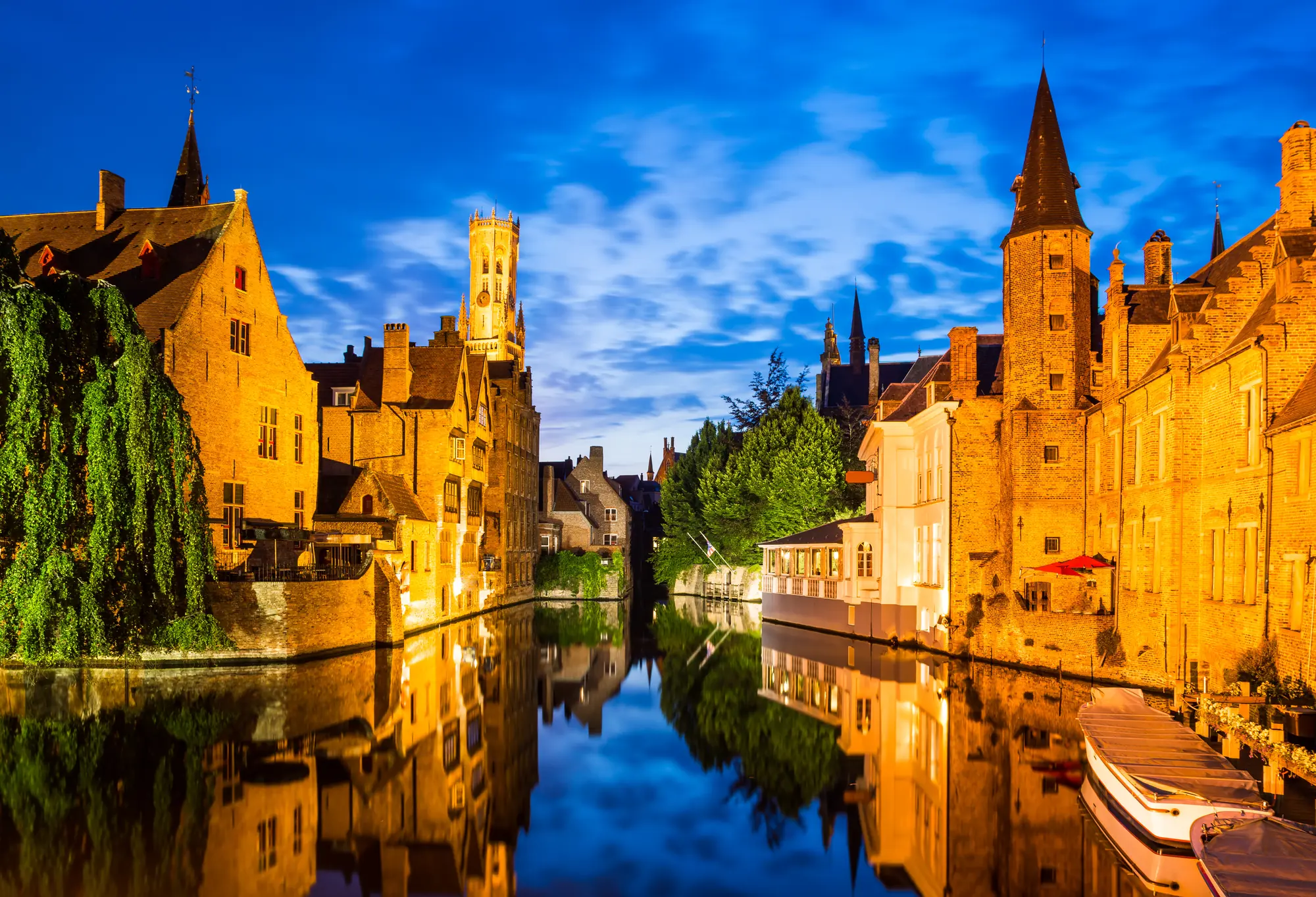 Illuminierter Rozenhoedkaai an einem milden Abend in Brügge, mit Beleuchtung und historischen Gebäuden, die sich sanft im Wasser spiegeln.