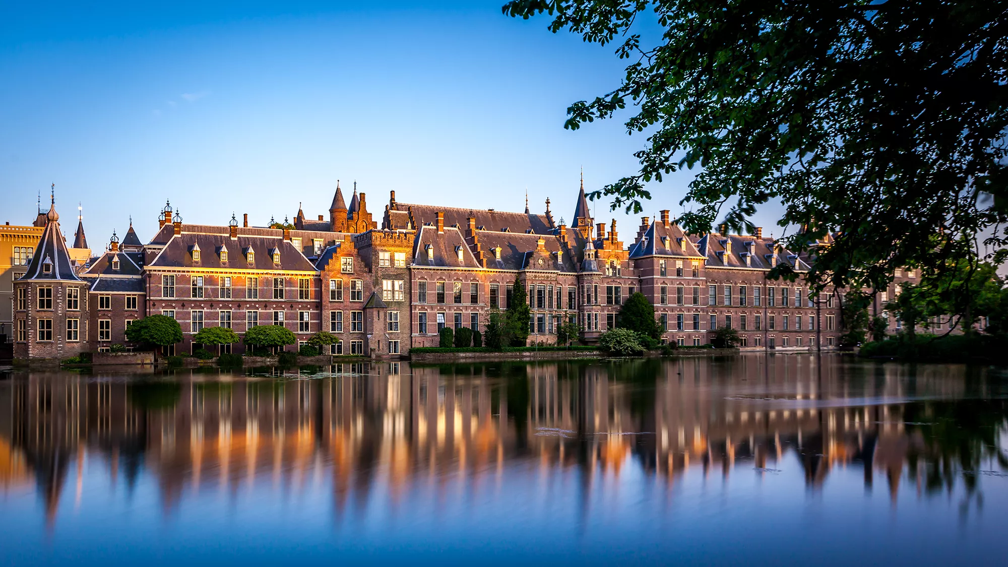 Ein Panorama des Binnenhofs in Den Haag, aufgenommen bei leichter Abenddämmerung. Sanfte Sonnenstrahlen reflektieren sich auf dem Wasser der Amstel und spiegeln die Architektur des historischen Gebäudes wider. Die ruhige Atmosphäre der Dämmerung verstärkt die Schönheit der Umgebung und lädt zur Besinnung ein.