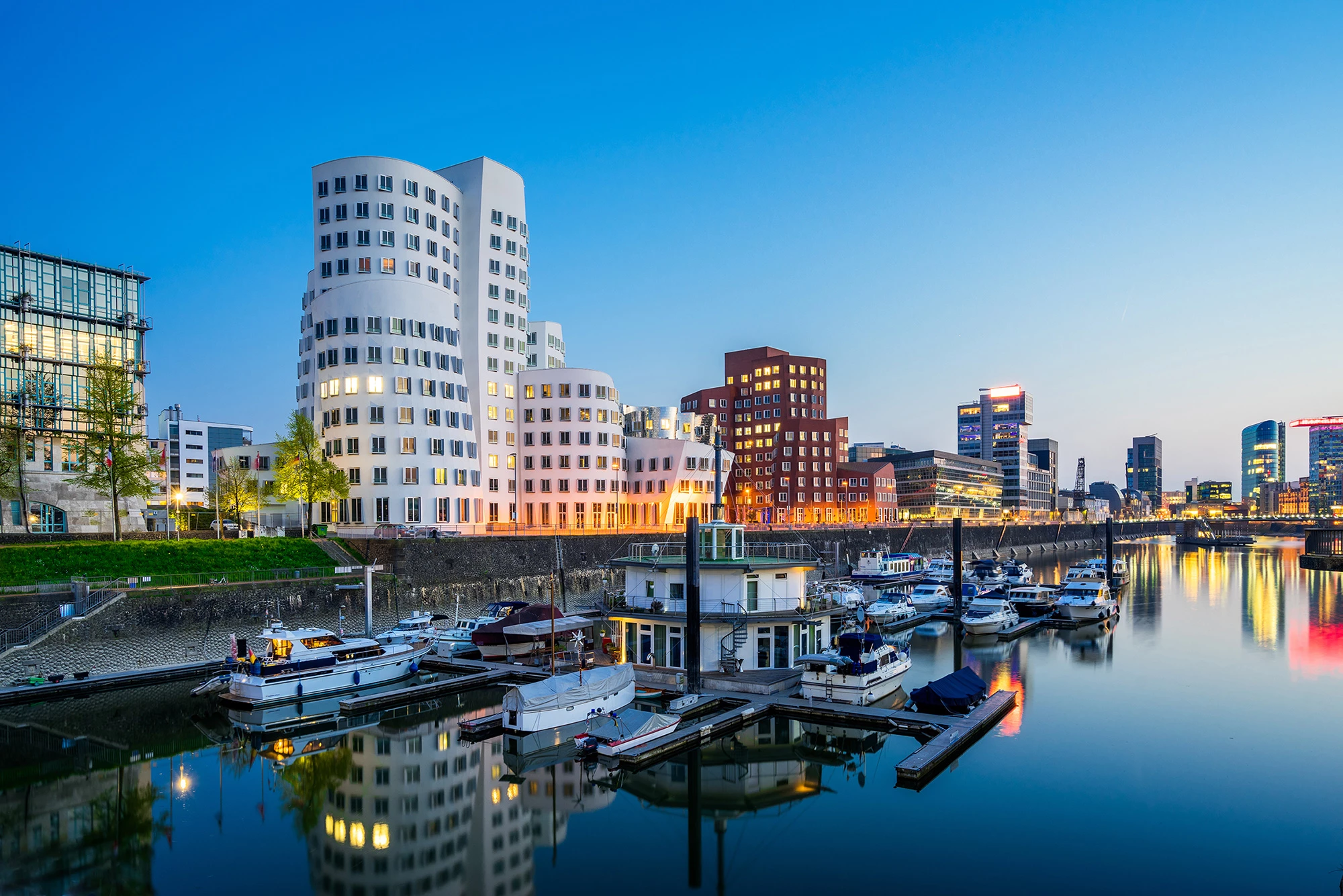 Abendliche Ansicht des Düsseldorfer MedienHafens mit modernen, architektonisch auffälligen Gebäuden, darunter die berühmten Gehry-Bauten. Im Vordergrund liegt der Hafen mit mehreren Booten, während sich die Gebäude und Lichter malerisch im Wasser spiegeln. Die Szenerie vereint moderne Architektur und urbanes Leben in einer ruhigen, maritimen Atmosphäre.