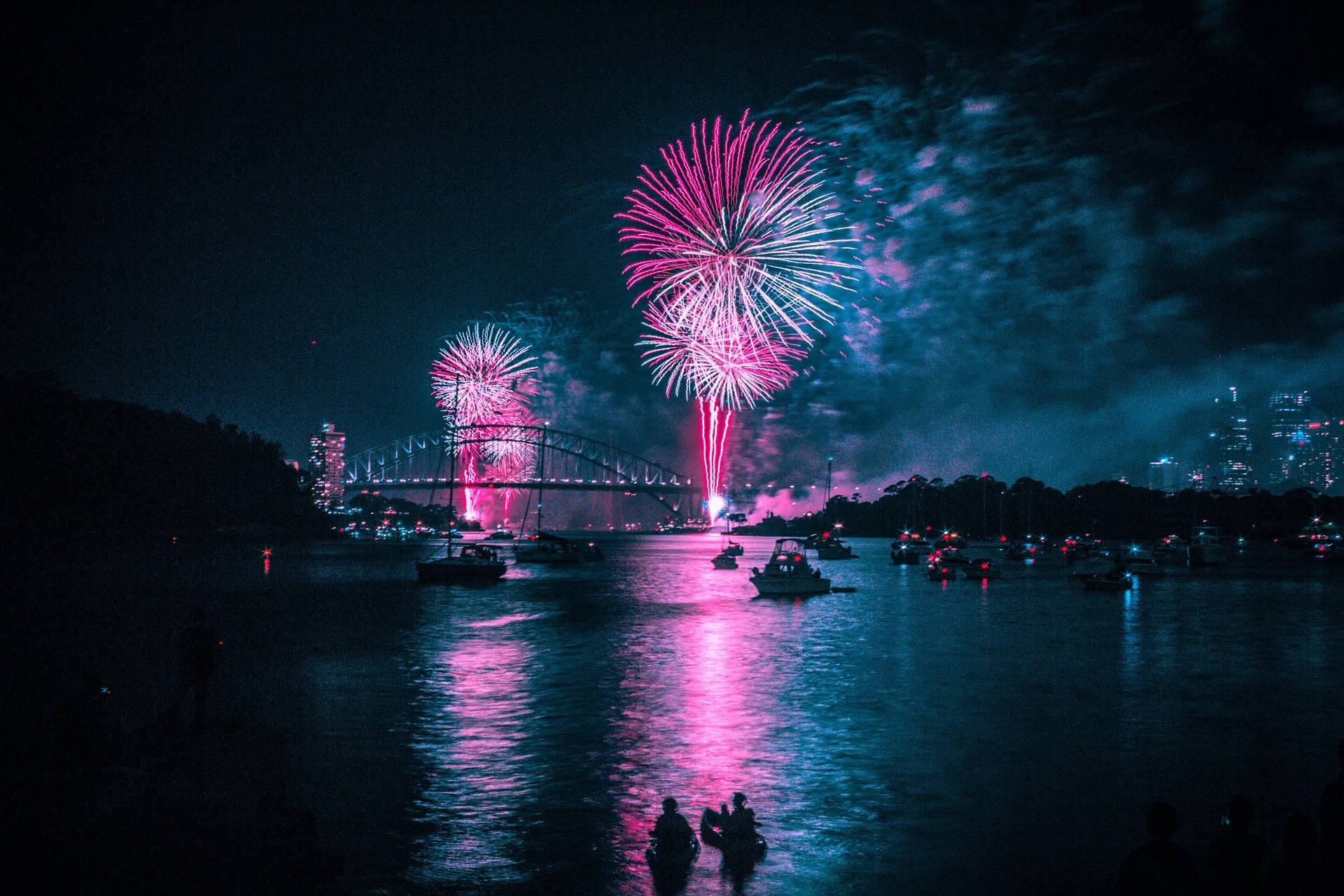 Nächtliches Feuerwerk über einer Flussbrücke. Auf dem Fluss sind Boote zu sehen und im Hintergrund die Skyline einer Stadt.