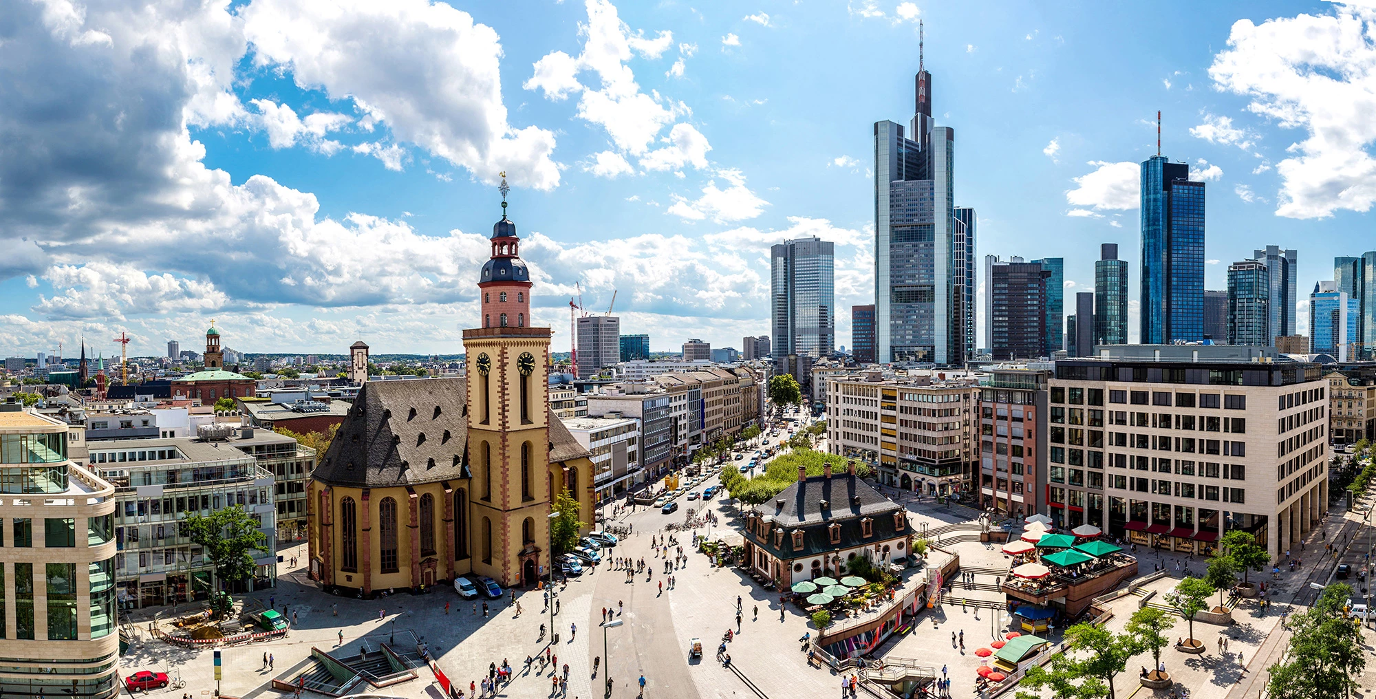 Luftaufnahme des Stadtzentrums von Frankfurt am Main mit Blick auf die Skyline. Im Vordergrund befindet sich die Katharinenkirche, umgeben von modernen Hochhäusern und urbanem Leben. Die Kombination aus historischer Architektur und modernen Wolkenkratzern zeigt das markante Stadtbild von Frankfurt am Main.