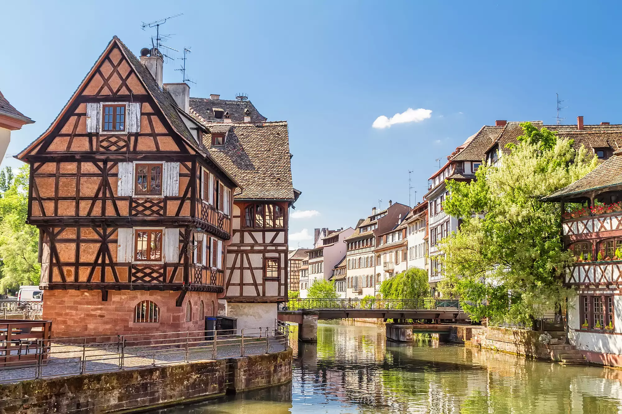 Fachwerkhäuserreihen in La Petite France bei sonnigem Wetter entlang des Flusses Ill, umgeben von grün leuchtenden Bäumen, schaffen eine einladende und malerische Atmosphäre.