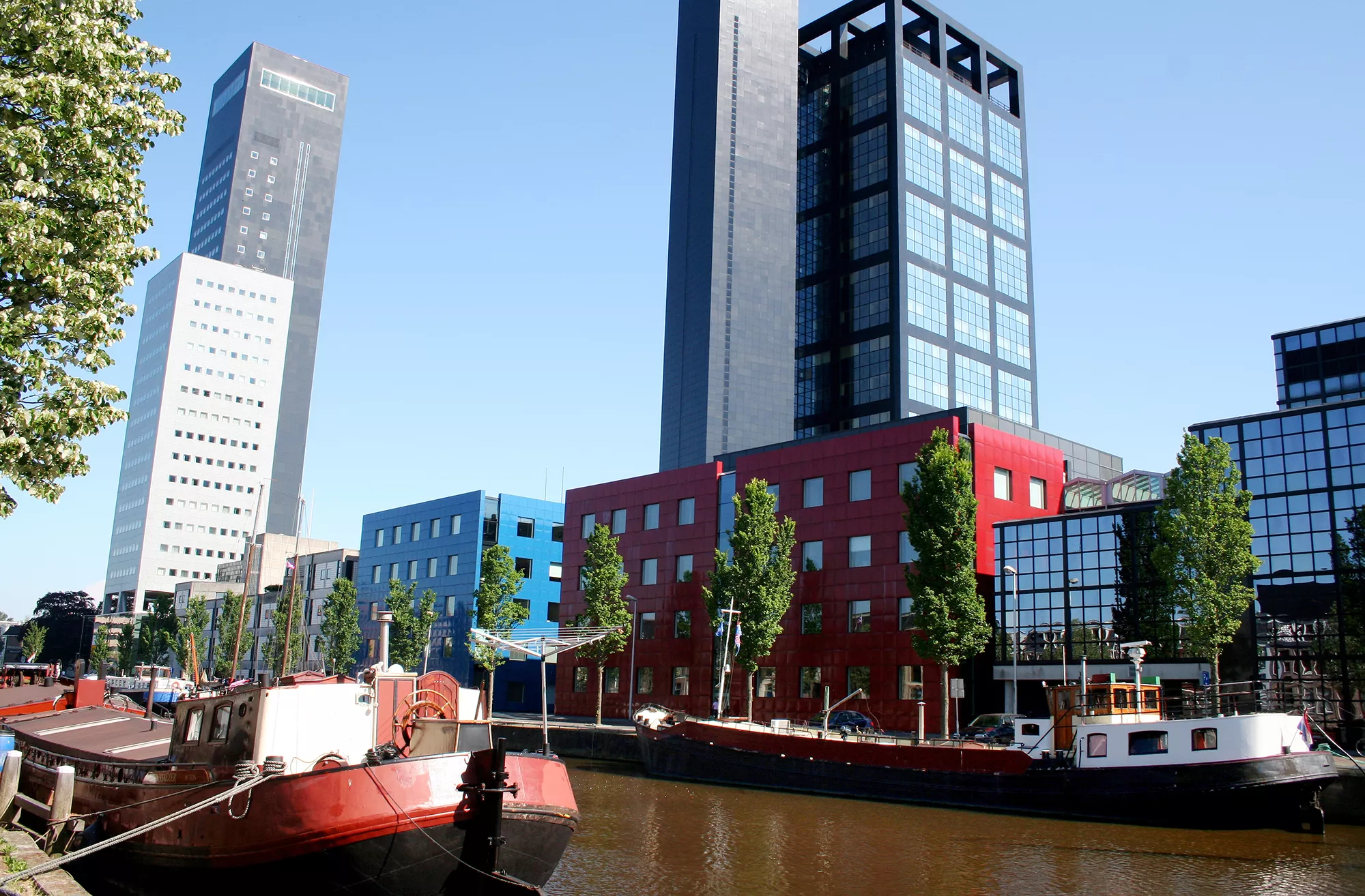 Der Achmea Turm in Leeuwarden, elegant am Canal Oldehove platziert, bei strahlendem Sonnenschein. Die klaren Linien des modernen Gebäudes harmonieren mit den angelegten Grachten, die die Umgebung umrahmen. Das sonnige Wetter bringt lebendige Farben hervor und unterstreicht die ansprechende Verbindung von zeitgenössischer Architektur und malerischer Wasserlandschaft.