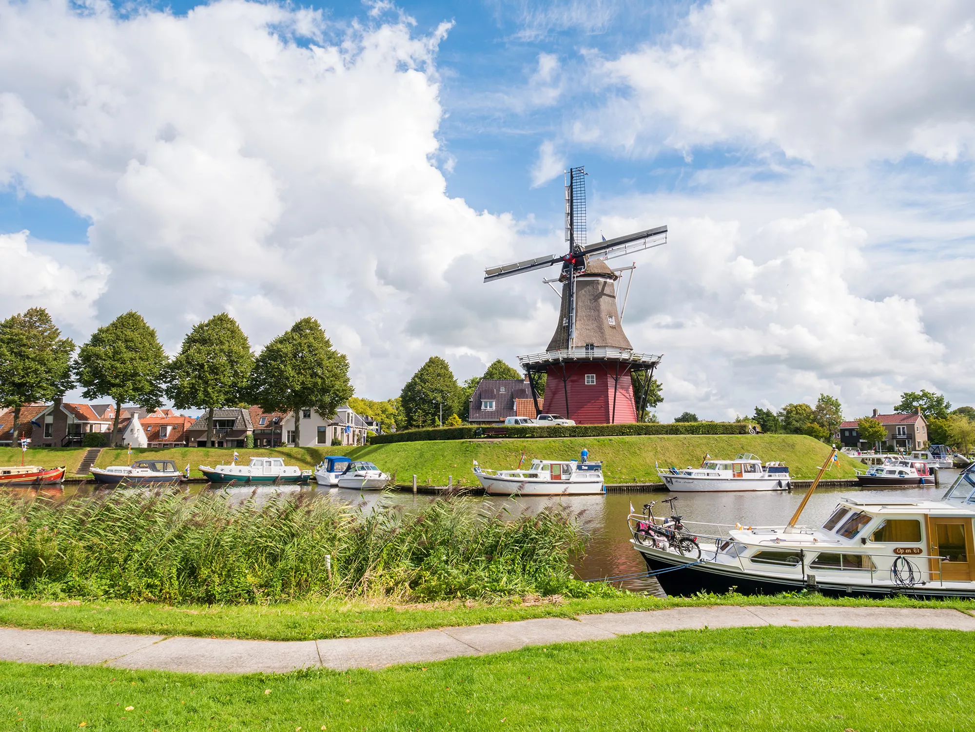 Die alte Windmühle Molen Zeldenrust am Kanal, umgeben von angelegten Booten und üppigem Grün in Friesland, bei Tageslicht. Die malerische Szenerie verbindet die traditionelle Architektur der Mühle mit der ruhigen Wasseroberfläche und der naturnahen Umgebung. Das Tageslicht hebt die Farben und Details der Mühle sowie der Flora hervor und schafft eine friedliche und einladende Atmosphäre.