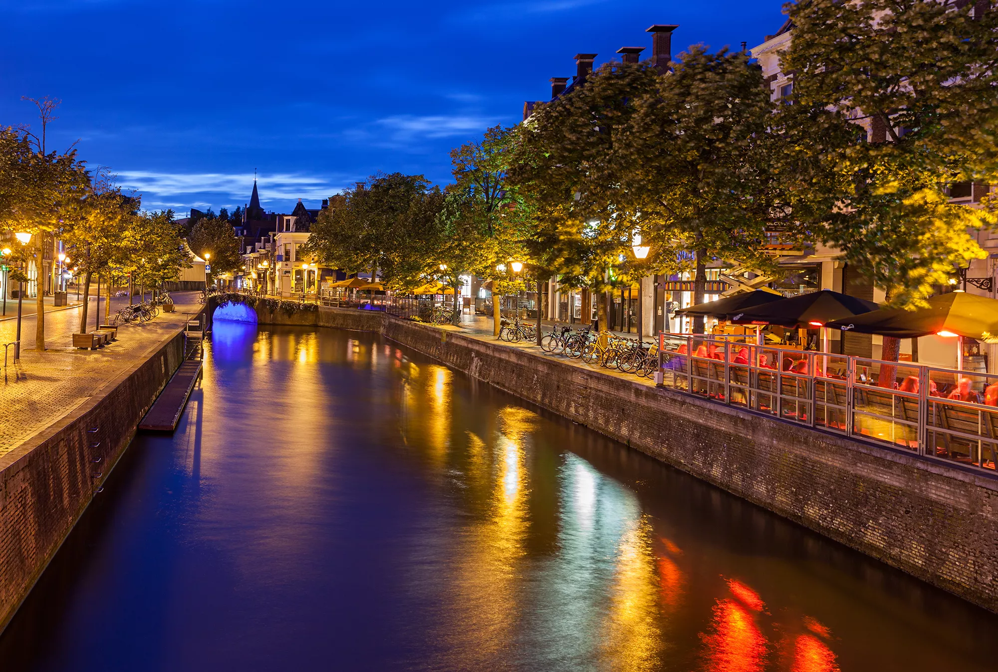 estaurants entlang eines beleuchteten Kanals in Leeuwarden am Abend. Die warmen Lichter reflektieren sich sanft im Wasser und schaffen eine einladende Atmosphäre. Die Architektur der Gebäude und die gemütlichen Terrassen laden Passanten zum Verweilen ein und vermitteln ein Gefühl von Geselligkeit und urbanem Leben in der malerischen Kulisse der Stadt.