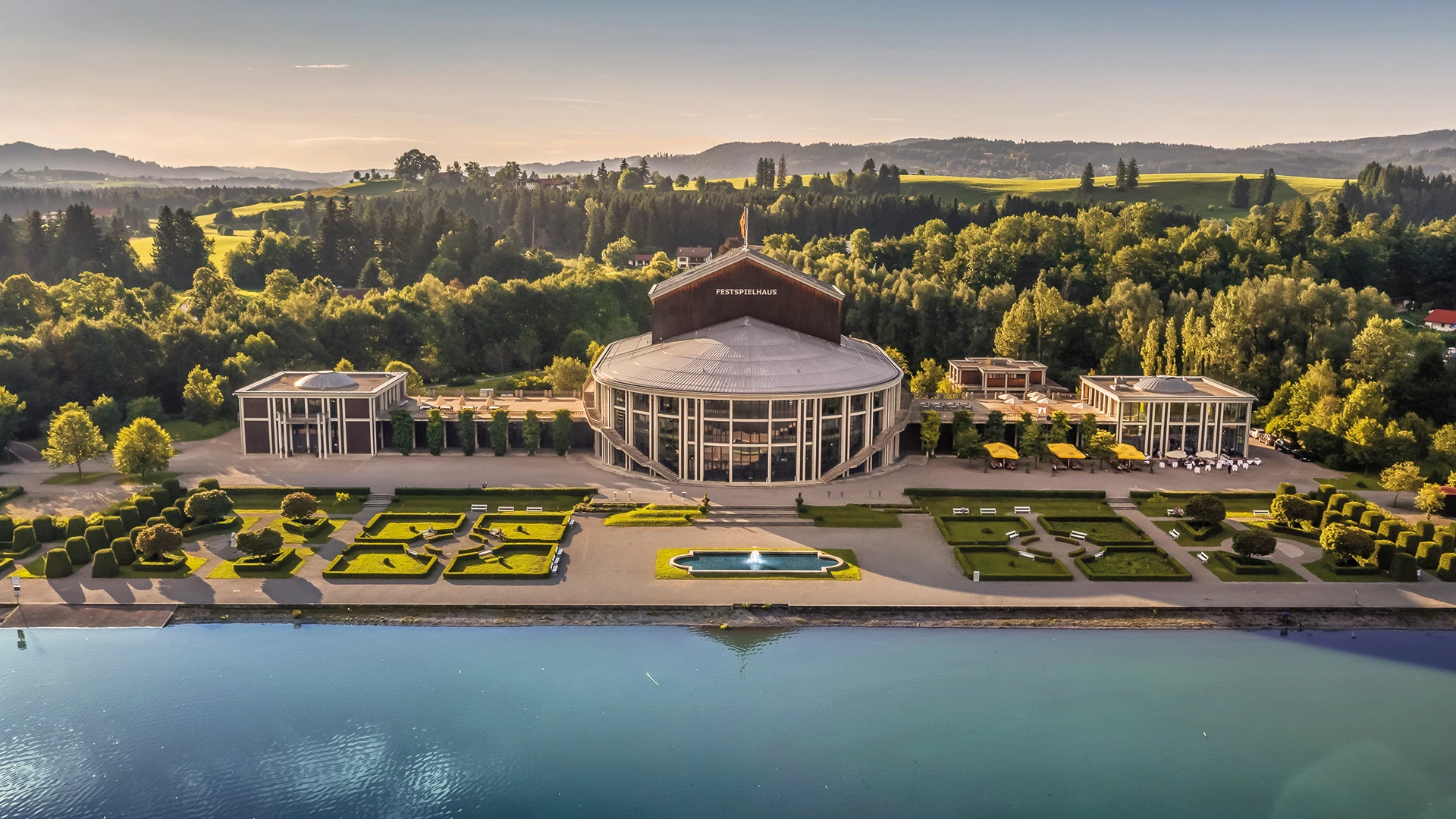 Das Bild zeigt das Festspielhaus Neuschwanstein aus der Vogelperspektive. Das Gebäude ist von gepflegten Gärten mit symmetrischen Grünflächen, einem Brunnen und Spazierwegen umgeben, die direkt an das blaue Ufer des Sees grenzen. Die idyllische Landschaft, eingerahmt von bewaldeten Hügeln, betont die harmonische Verbindung zwischen der modernen Architektur des Festspielhauses und der umgebenden Natur, was dem Ort eine besondere Eleganz verleiht.