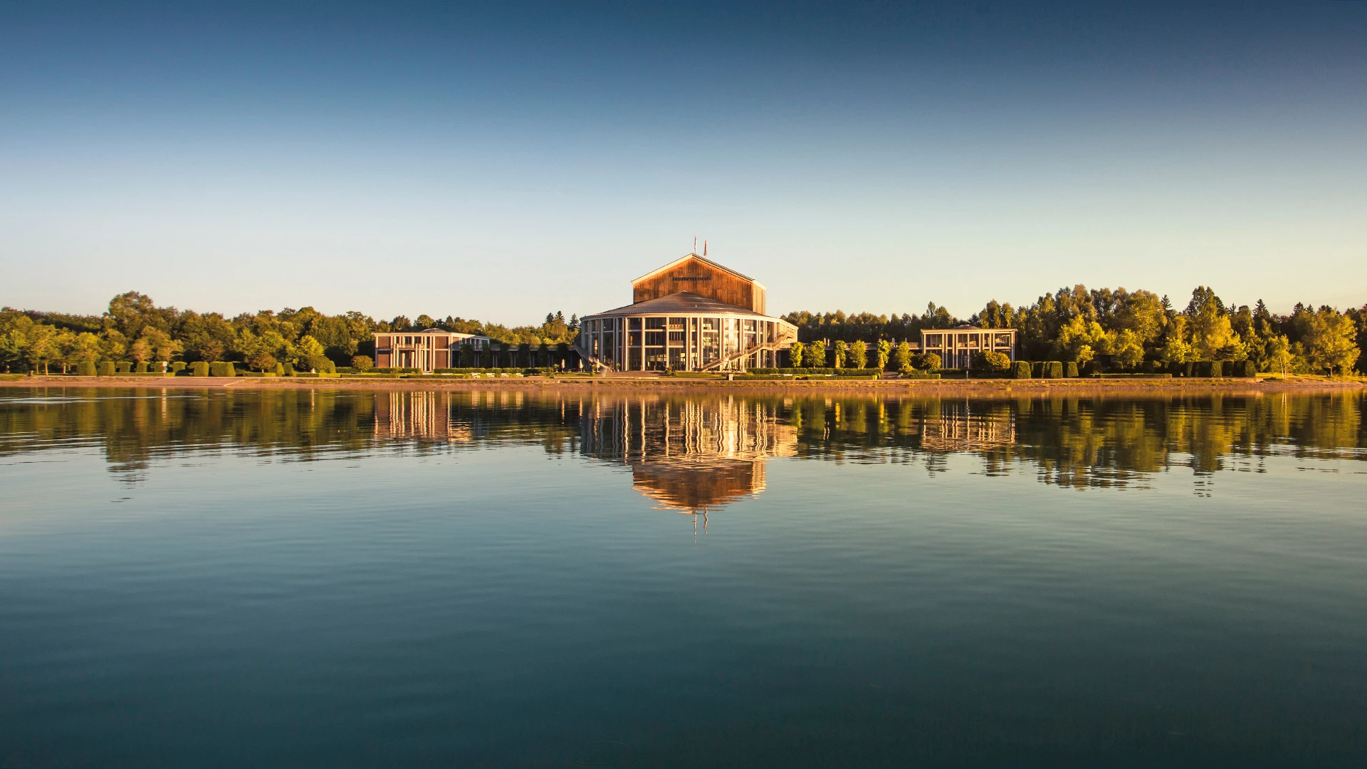 Das Bild zeigt das Festspielhaus Neuschwanstein, idyllisch am Ufer eines Sees gelegen. Das Gebäude spiegelt sich perfekt in der ruhigen Wasseroberfläche, während die umgebende Natur in warmen Sonnenlicht getaucht ist. Die harmonische Verbindung von moderner Architektur und malerischer Landschaft verleiht dem Festspielhaus eine besonders eindrucksvolle und friedliche Ausstrahlung.