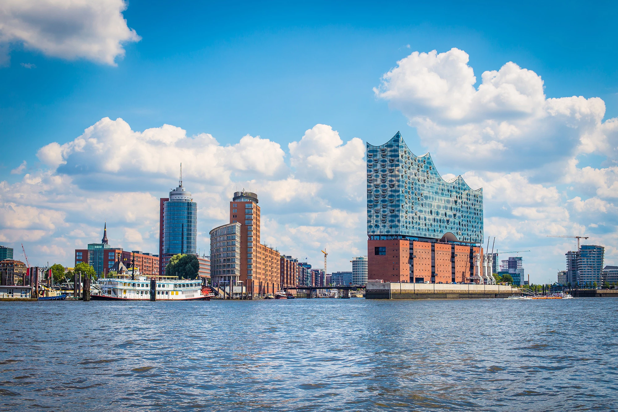 Die Elbphilharmonie in Hamburg, ein beeindruckendes Wahrzeichen der Stadt, thront mit ihrer wellenförmigen Glasfassade über dem historischen Hafengebäude in der Höhe. Im Vordergrund das Wasser der Elbe, das die moderne Architektur und den klaren blauen Himmel reflektierten.