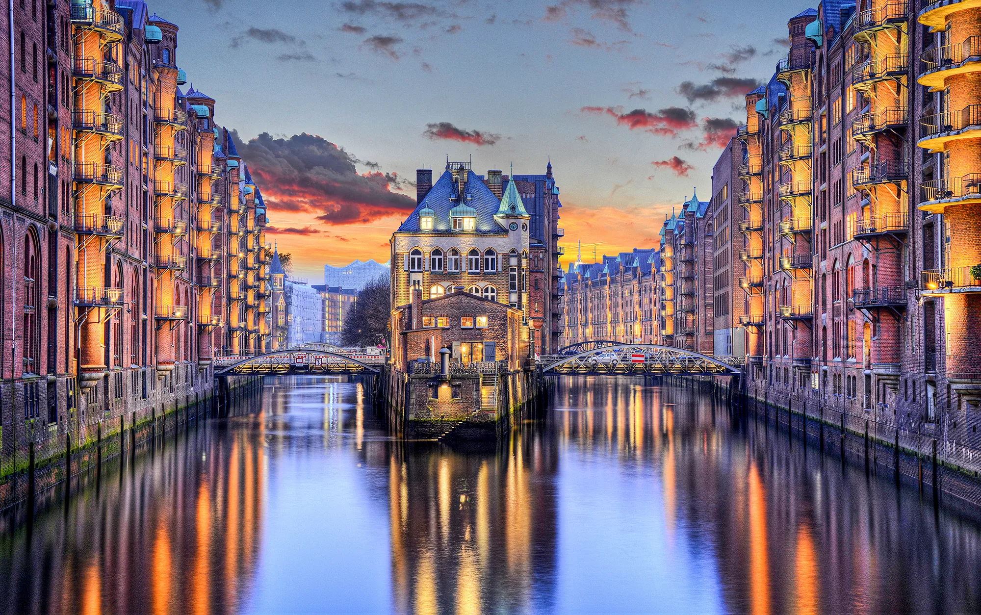Das Bild zeigt die Hamburger Speicherstadt in der Dämmerung. Die historischen Backsteingebäude, die sich entlang der Kanäle erstrecken, sind stimmungsvoll beleuchtet und spiegeln sich im ruhigen Wasser wider. Im Zentrum des Bildes befindet sich das markante Wasserschloss, das von zwei Brücken umgeben ist. Im Hintergrund ist die moderne Architektur der Elbphilharmonie zu erkennen, die einen Kontrast zur traditionellen Speicherstadt bildet.