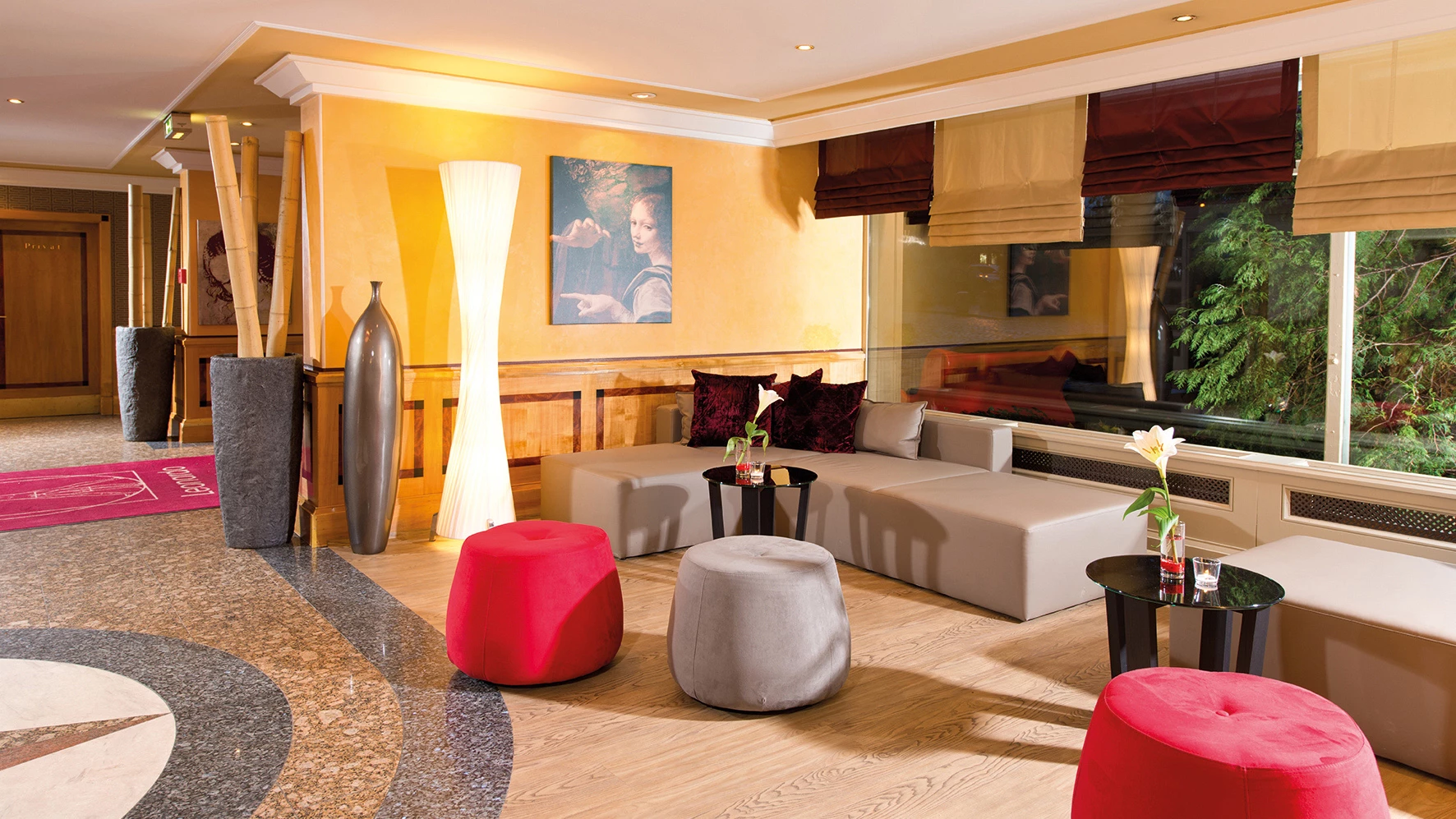 Gemütliche Hotellobby mit grauer Eckcouch, roten und grauen Sitzhockern, kleinen schwarzen Tischen mit Blumen und großen Vasen neben einer Stehlampe