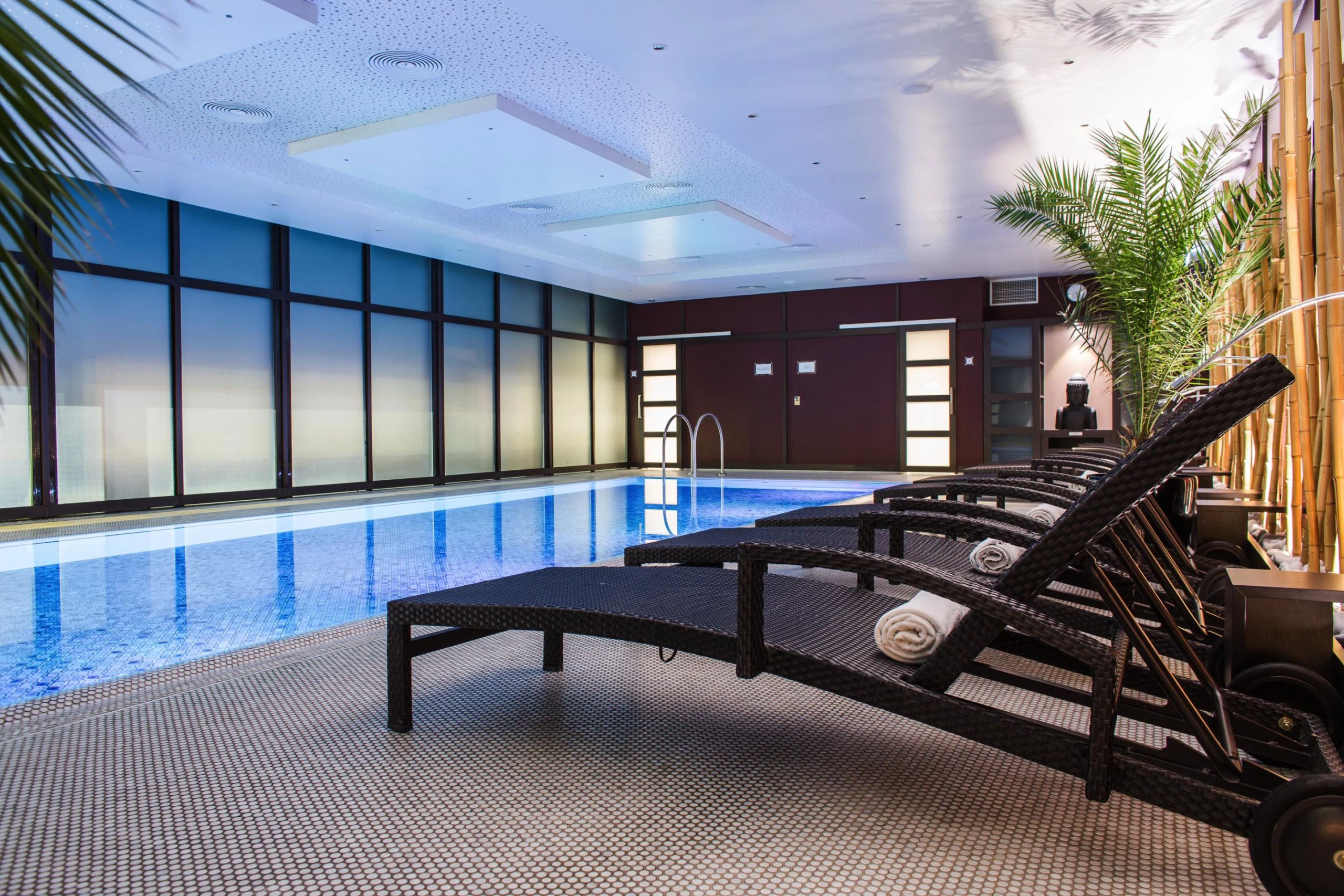 Eleganter Innenpool im Steigenberger Graf Zeppelin Stuttgart mit modernen Liegestühlen und dezenter Beleuchtung. Das klare, blaue Wasser des Pools und die stilvolle Dekoration mit Bambus und Palmen sorgen für eine entspannte Atmosphäre.