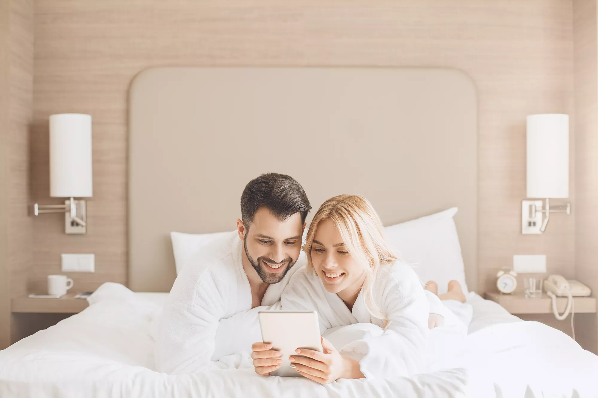 Zimmerbeispiel, junges fröhliches Paar liegt auf dem Bett und sieht in ein Tablet