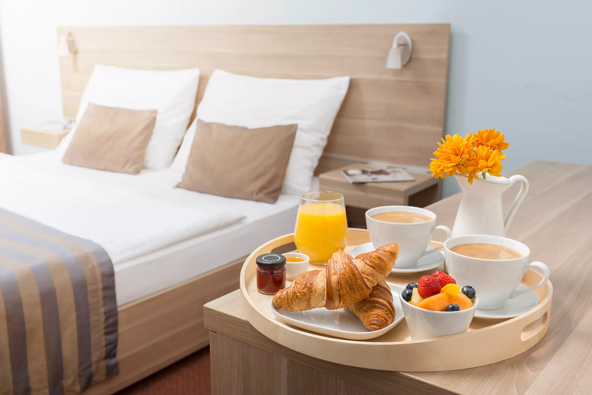Zimmerbeispiel Doppelzimmer Neben dem Bett auf den Tisch ein Frühstückstablett mit 2 Tassen Kaffee, Croissants, Marmelade, Schale Obst und Orangensaft.