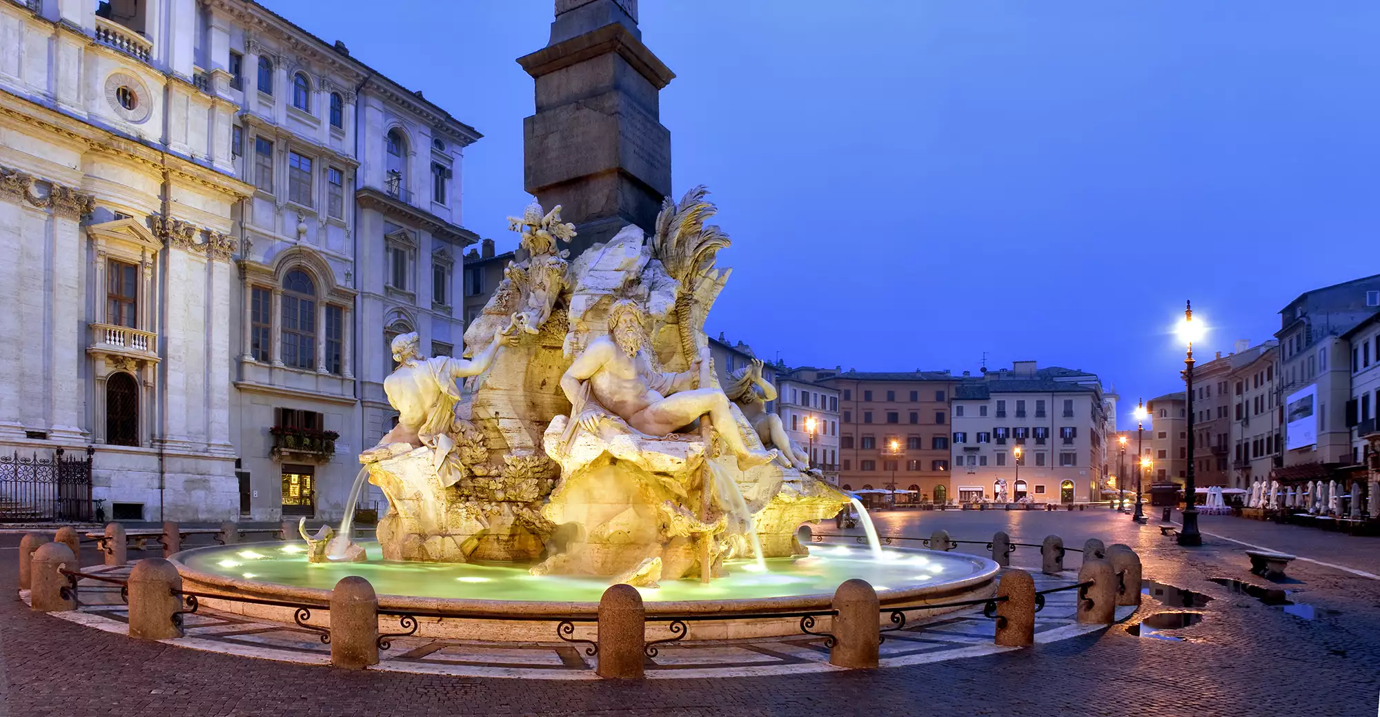 Nahaufnahme des Piazza Navona am Abend, beleuchtet von stimmungsvoller Stadtbeleuchtung. Die beeindruckenden Brunnen und historischen Fassaden strahlen im warmen Licht, während die ruhige Atmosphäre des Platzes durch die sanfte Beleuchtung und die architektonischen Details verstärkt wird.