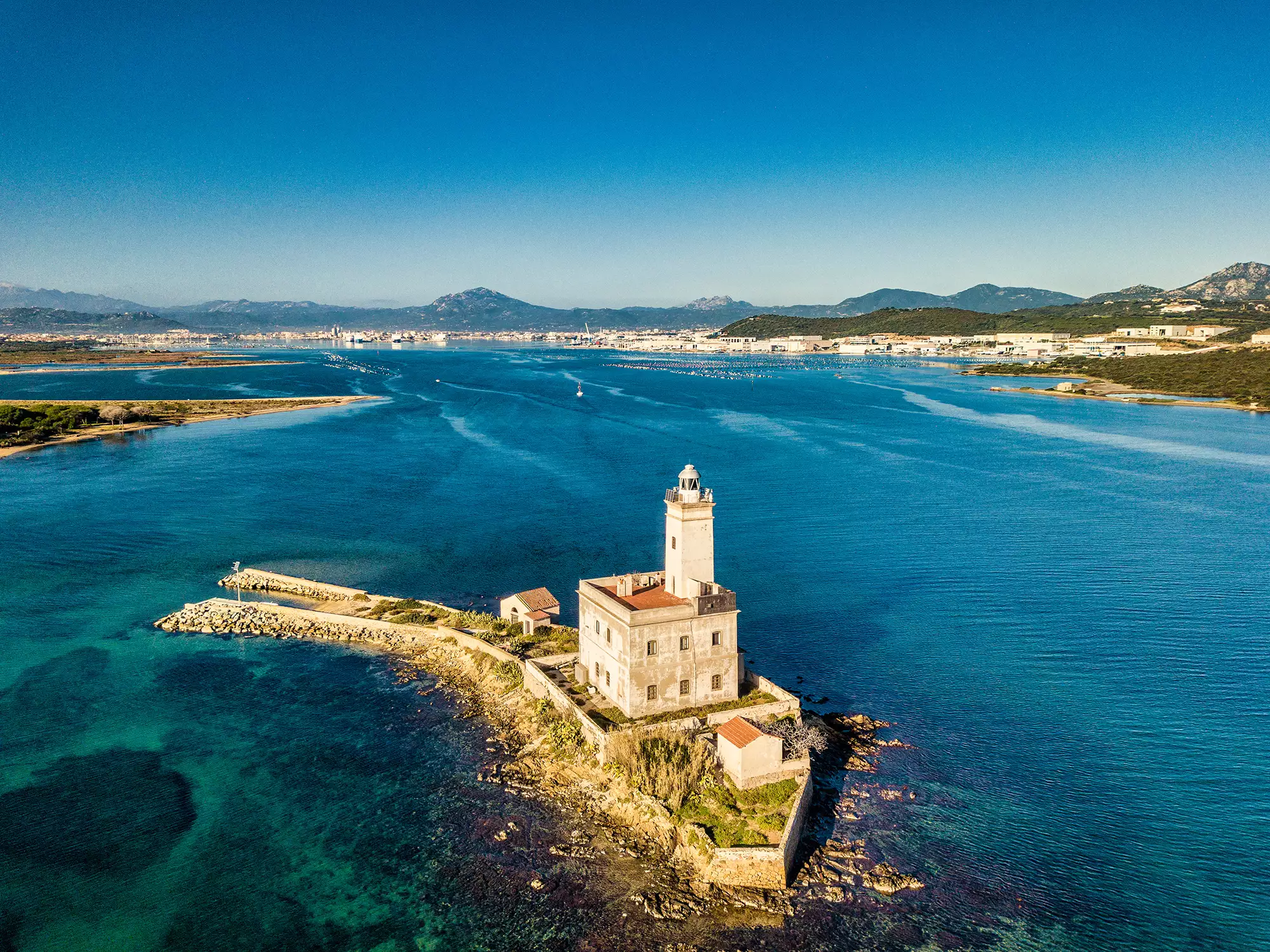 Panoramaansicht von Olbia, das die Hafeneinfahrt mit dem schönen blauen Meer zeigt. In der Ferne sind kleine Schiffe zu sehen, während die umgebende Natur mit sanften Bergen die Szenerie umrahmt. Das helle Tageslicht verstärkt die Farben und schafft eine einladende, idyllische Atmosphäre.