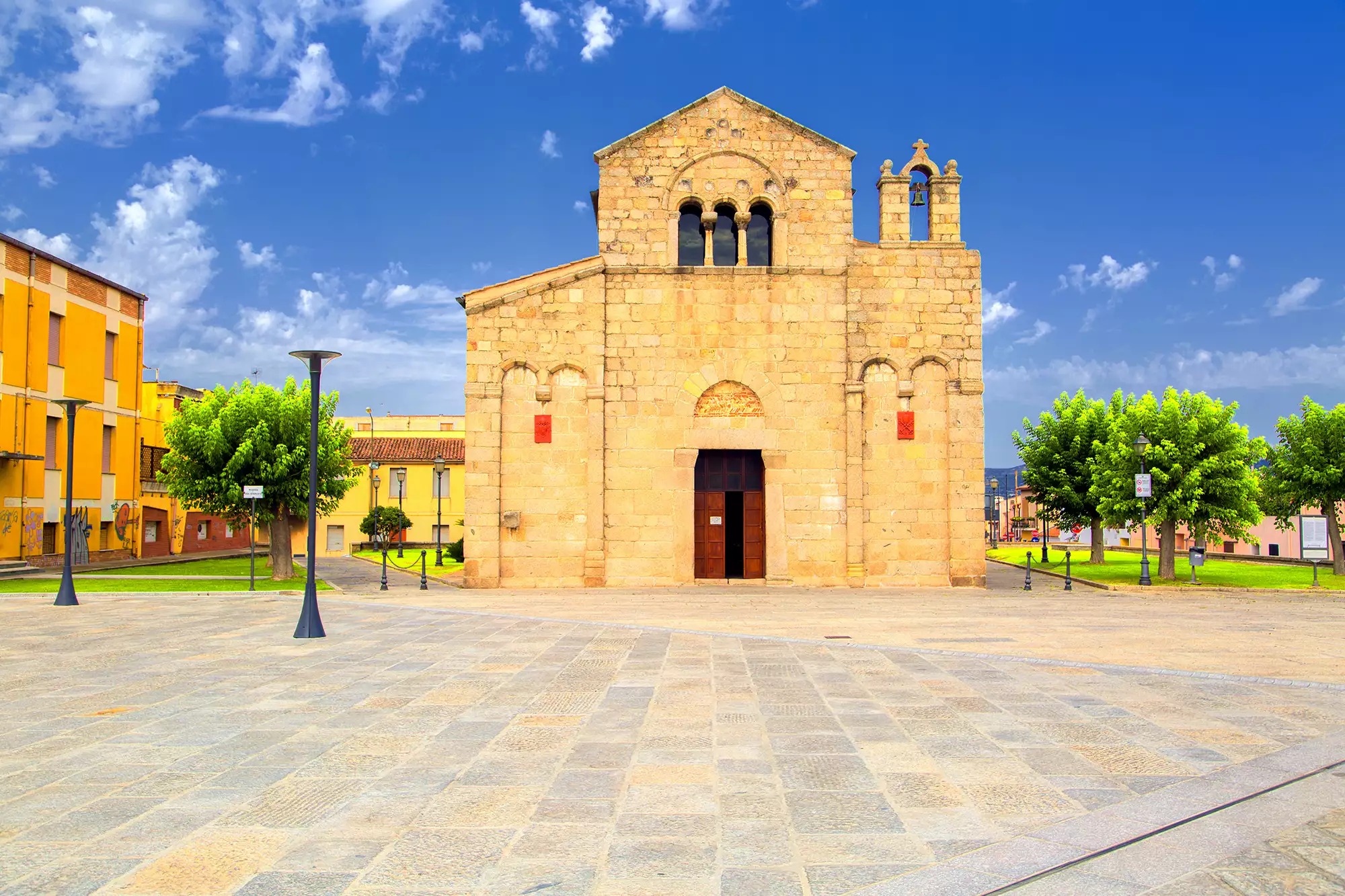 San Simplicio in Olbia bei sonnigem Tageslicht. Die beeindruckende Fassade der Kirche strahlt in warmen Farben und wird von der klaren, blauen Himmelkulisse ergänzt. Die historischen Details und die umgebende Architektur betonen die kulturelle Bedeutung dieses Wahrzeichens und schaffen eine einladende Atmosphäre.