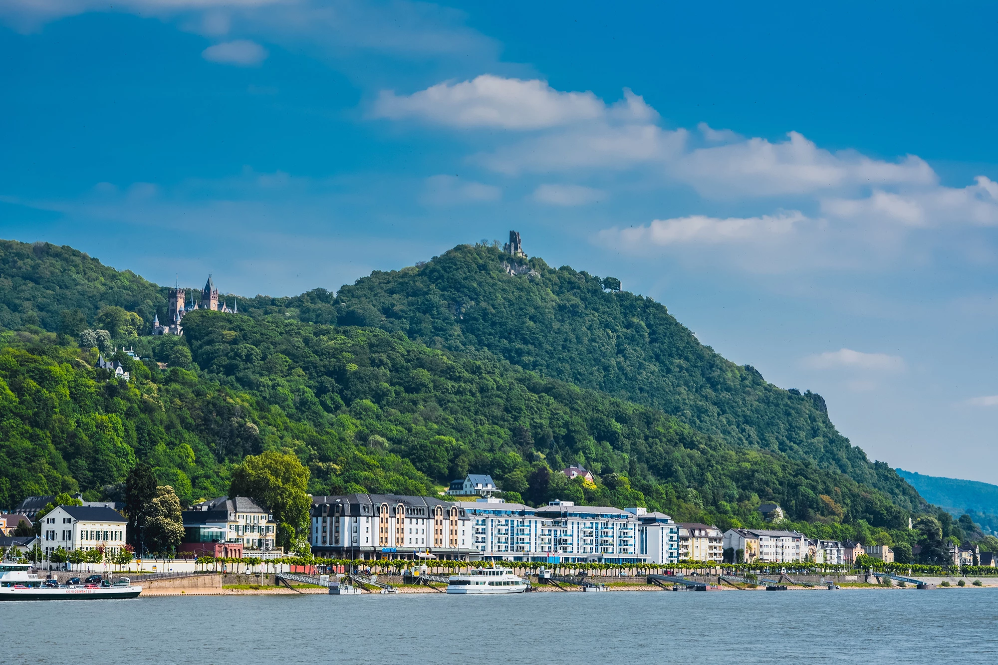 Das Stadtmotiv von Königswinter zeigt die malerische Stadt am Rhein, eingebettet in grüne üppige Hügellandschaften. Im Hintergrund erhebt sich der Petersberg mit der Burgruine Drachenfels, während die prächtigen Gebäude und Hotels entlang der Rheinpromenade die Uferlinie säumen. Die Szenerie wird durch das ruhige, blaue Wasser des Rheins ergänzt, über das ein Ausflugsschiff gleitet, und verleiht dem Bild eine idyllische und entspannte Atmosphäre.