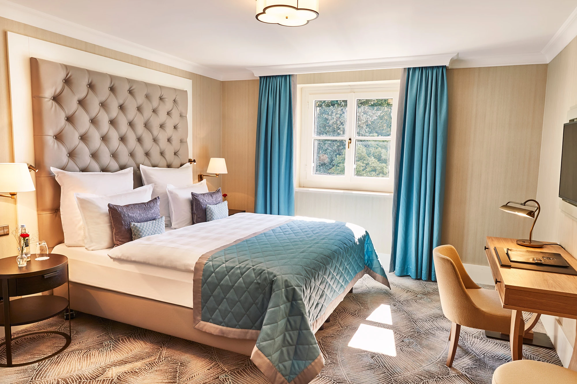 Das Bild zeigt ein elegantes Beispielzimmer im Steigenberger Hotel Petersberg. Das Zimmer besticht durch ein luxuriöses Design, das mit sanften Beigetönen und türkisfarbenen Akzenten harmoniert. Das große, gepolsterte Kopfteil des Bettes verleiht dem Raum eine edle Note. Neben dem gemütlichen Bett befinden sich moderne Nachttische und Lampen, während ein stilvoller Schreibtisch mit Leselampe zum Arbeiten einlädt. Die großen Fenster lassen viel Tageslicht herein und bieten einen Blick ins Grüne, was eine ruhige und entspannende Atmosphäre schafft.