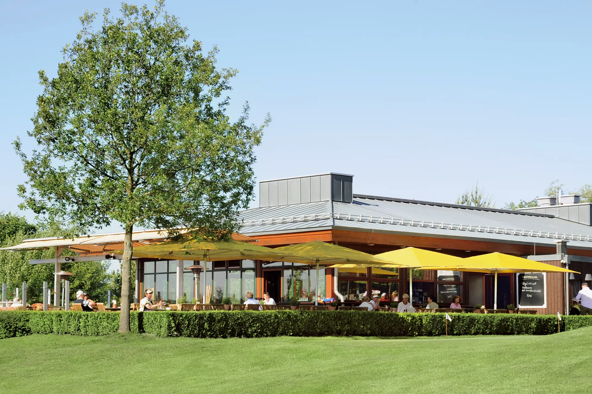 Blick auf die Terrasse des Golfclub des Lindner Hotel & Sporting Club Wiesensee eingerichtet mit Sitzgruppen unter schattenspendenden Sonnenschirmen