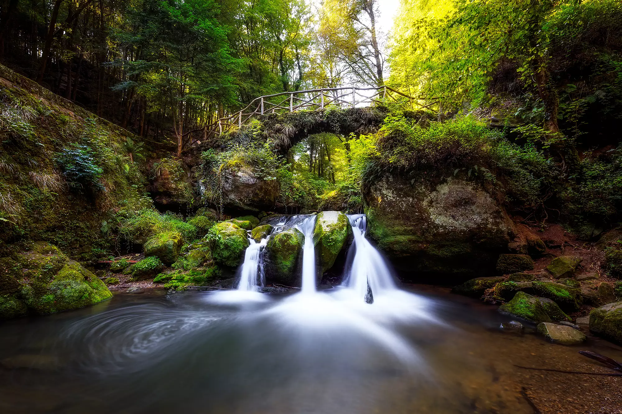 Illustration des Wasserfalls am Schiessentümpel im Müllerthal, umgeben von einer bewachsenen Brücke. Die grüne Vegetation und das plätschernde Wasser schaffen eine idyllische und entspannende Atmosphäre.