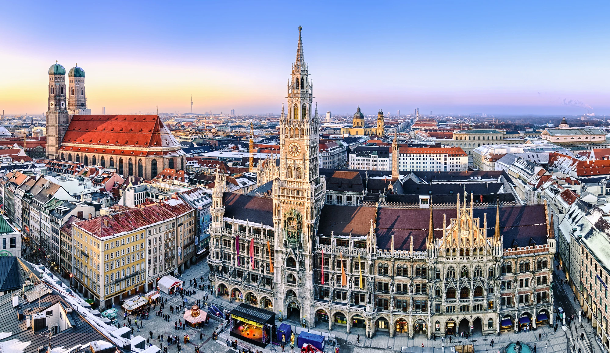 Das Bild zeigt ein beeindruckendes Panorama der Münchener Innenstadt mit dem Neuen Rathaus am Marienplatz im Zentrum. Die gotische Architektur des Rathauses mit seinem markanten Turm und das lebendige Treiben auf dem Platz fügen sich harmonisch in das Stadtbild ein. Im Hintergrund erhebt sich die imposante Frauenkirche mit ihren markanten Zwiebeltürmen, während die malerischen Dachlandschaften Münchens in den goldenen Schein des Abendlichts getaucht sind.