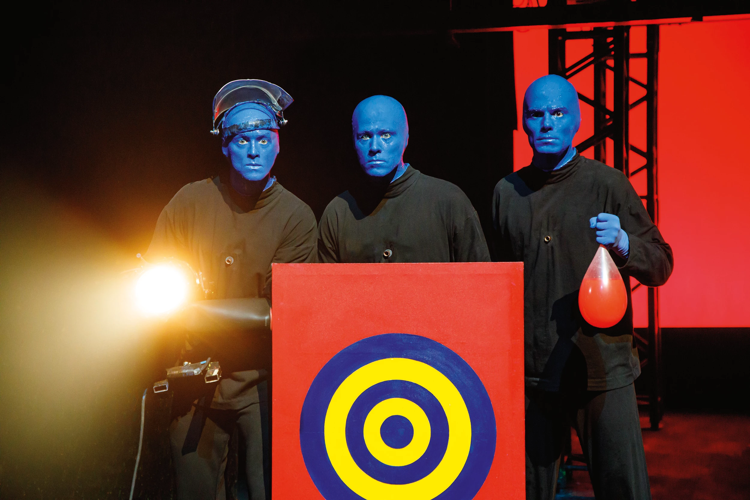 BLUE MAN GROUP Show in Berlin -Szene 3 blaue Männer in schwarzer Kleidung stehen mit einer Lampe und einer Zielscheibe auf der Bühne